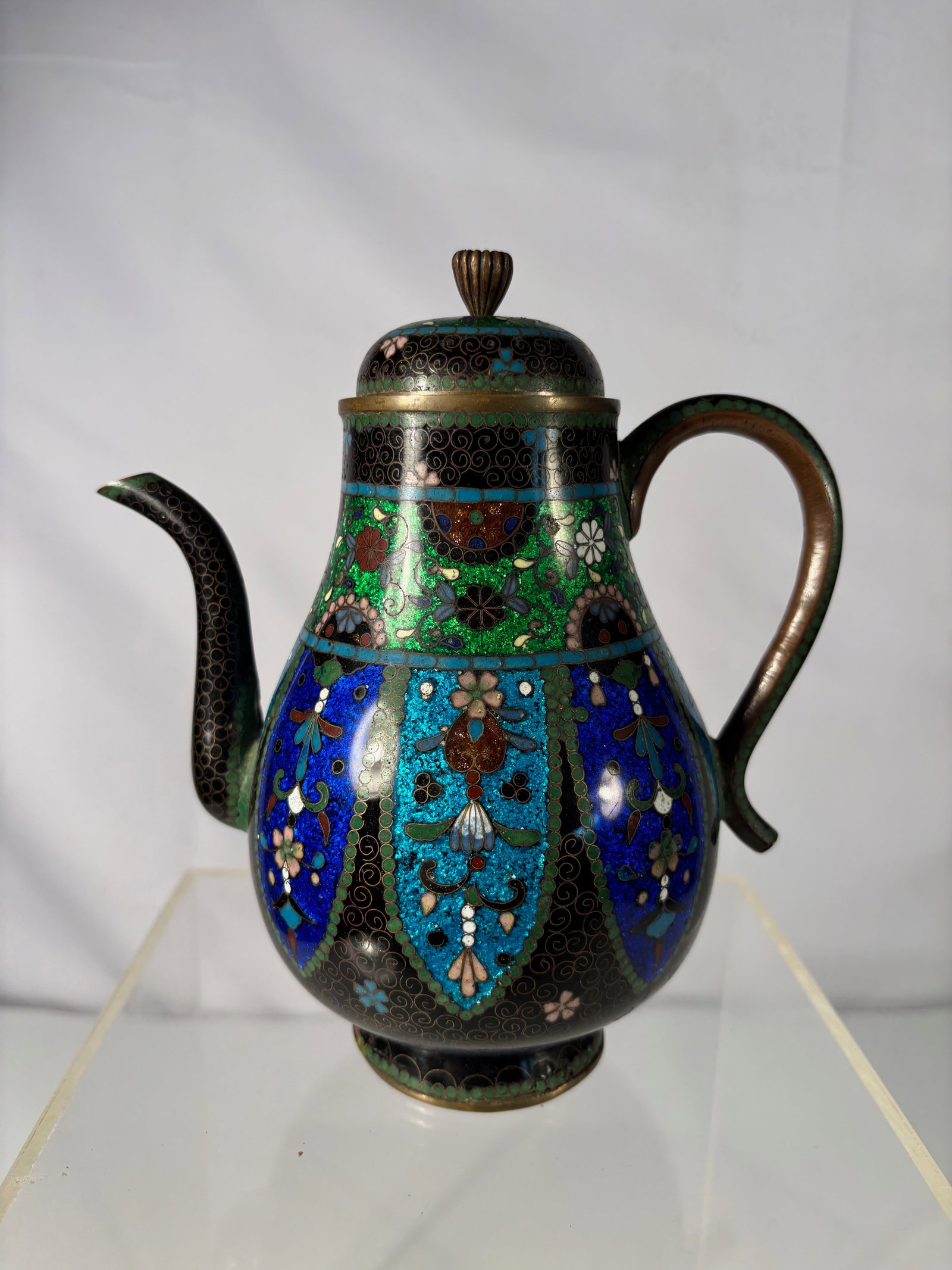 A Japanese Cloisonné Enamel Teapot: Meiji period(1868-1912) , of pear-shaped form with domed lid and gilt-metal finial, decorated with alternating panels of floral motifs in cobalt blue, turquoise, green, and aubergine enamels on a blac