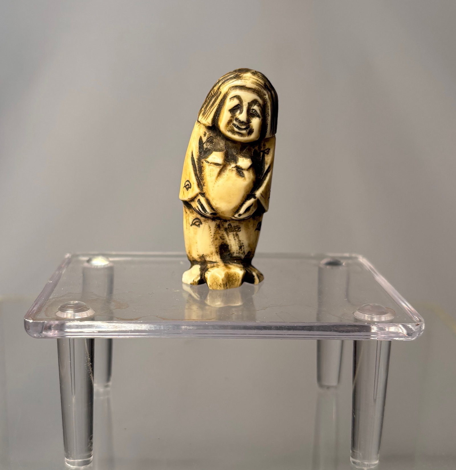 A Japanese Figural Netsuke, Carved Bone Standing Figure, Signed (Hisakazu / Ky?ichi), Meiji period: 19th century, A Japanese figural netsuke carved from bone, depicting a standing human figure with a humorous expression and stylized features. The carving includes two himotoshi to the reverse,