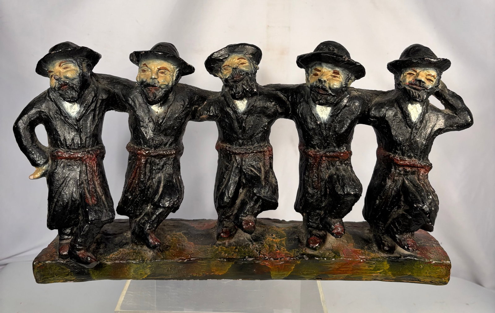 Painted Pottery Figural Group — Dancing Hasidic/Klezmer Figures: A polychrome painted pottery figural group depicting five bearded men in traditional dark coats and hats, arms linked in a dancing line, painted in black with cream/flesh-toned faces and hands, mounte