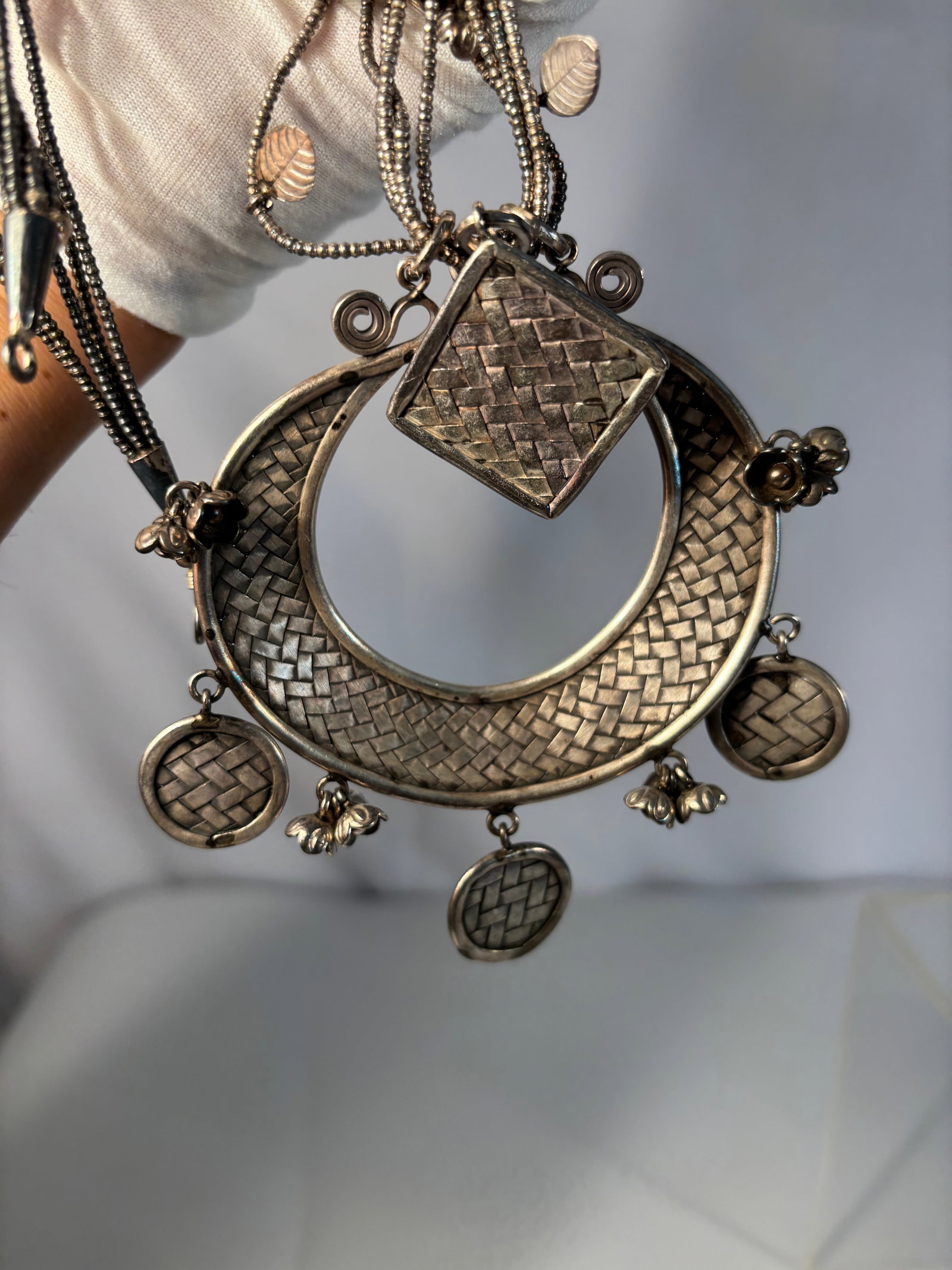 Silver Multi-Strand Necklace with Large Crescent Pendant and Decorative Disc Charms: Striking necklace featuring multiple silver chains adorned with small leaf and bead accents, suspending a large crescent-shaped pendant with woven-pattern surface and a central diamond-shaped element.