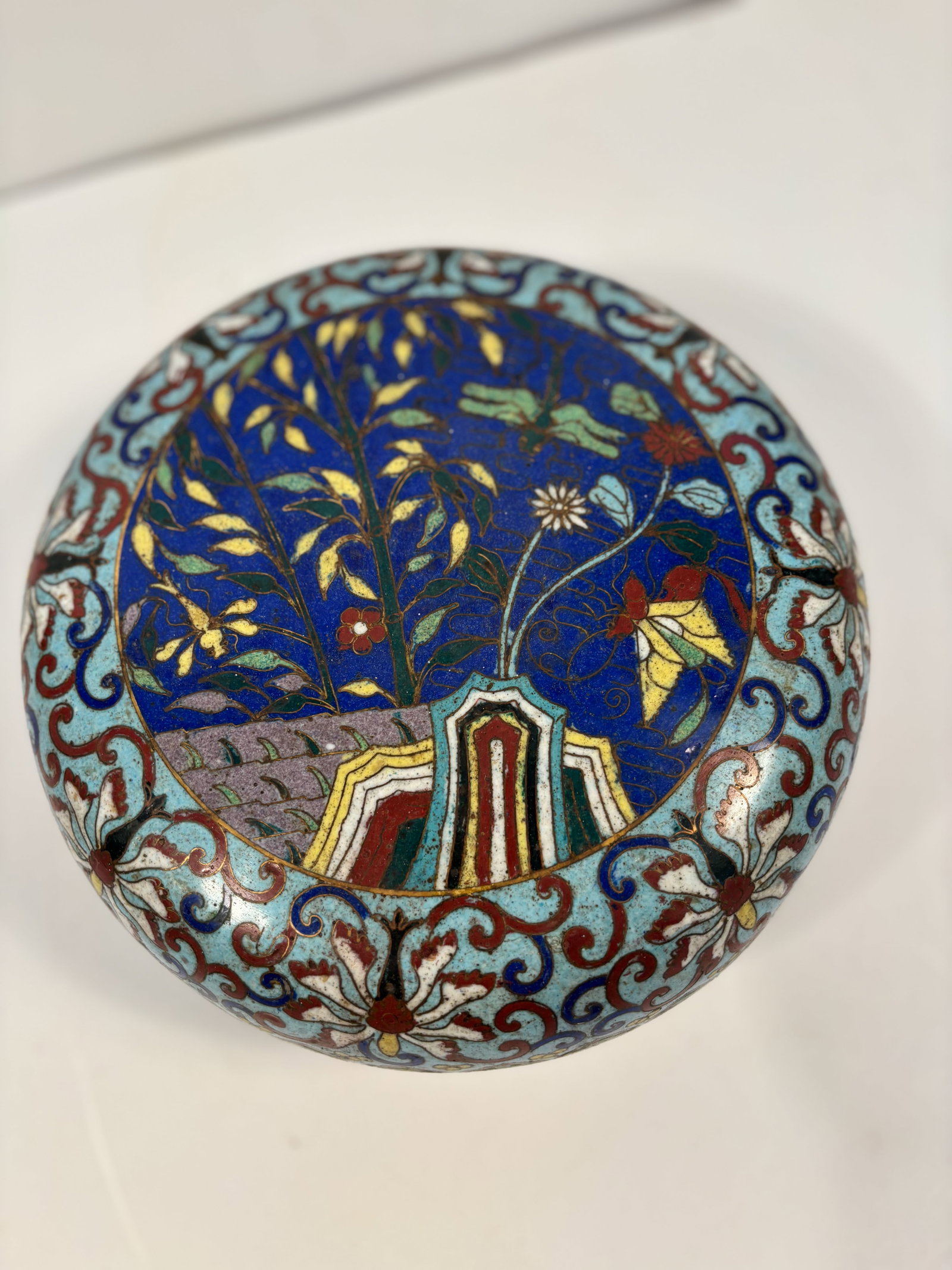 A Chinese cloisonné enamel box: 18th -19th century; The top shows a garden scene with a tree and architecture,Surrounding floral motifs ,The border features Ruyi head. Dimensions?Diameter: 8.5 inches; Height: 4 inches.