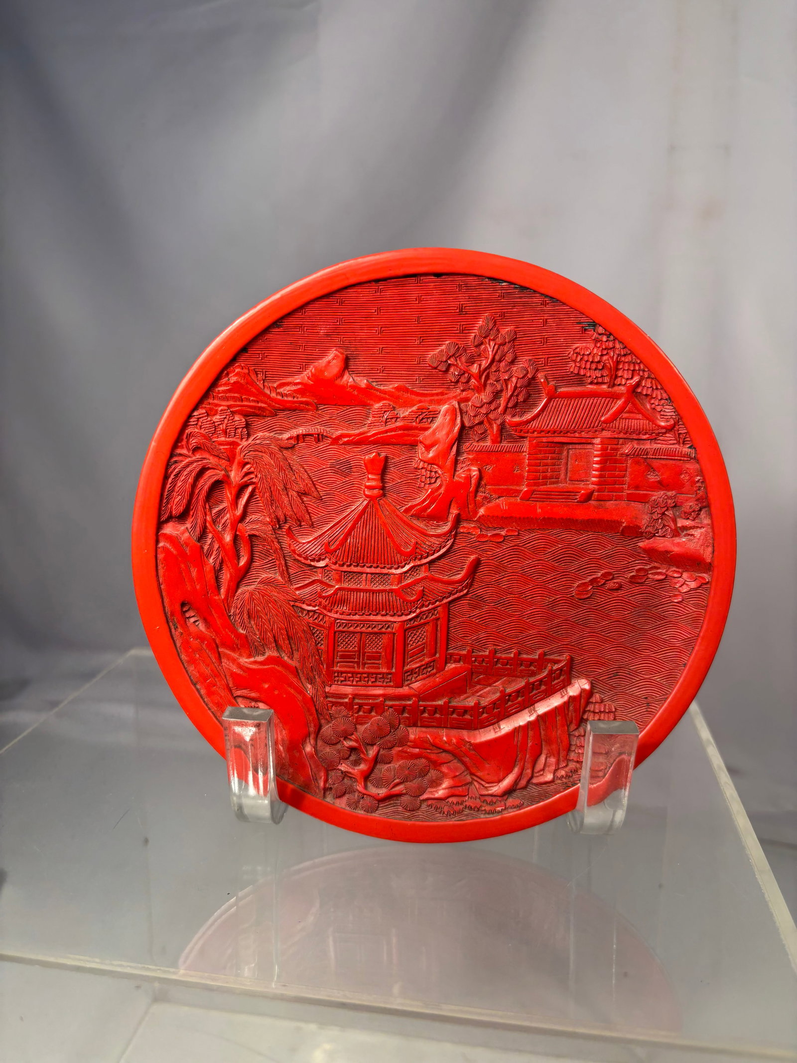 A Chinese Carved Cinnabar-Style Lacquer Landscape Plate: Chinese carved cinnabar-style decorative plate depicting a traditional pavilion overlooking a river landscape with bridges, trees, mountains, and architectural structures. Executed in relief carving w