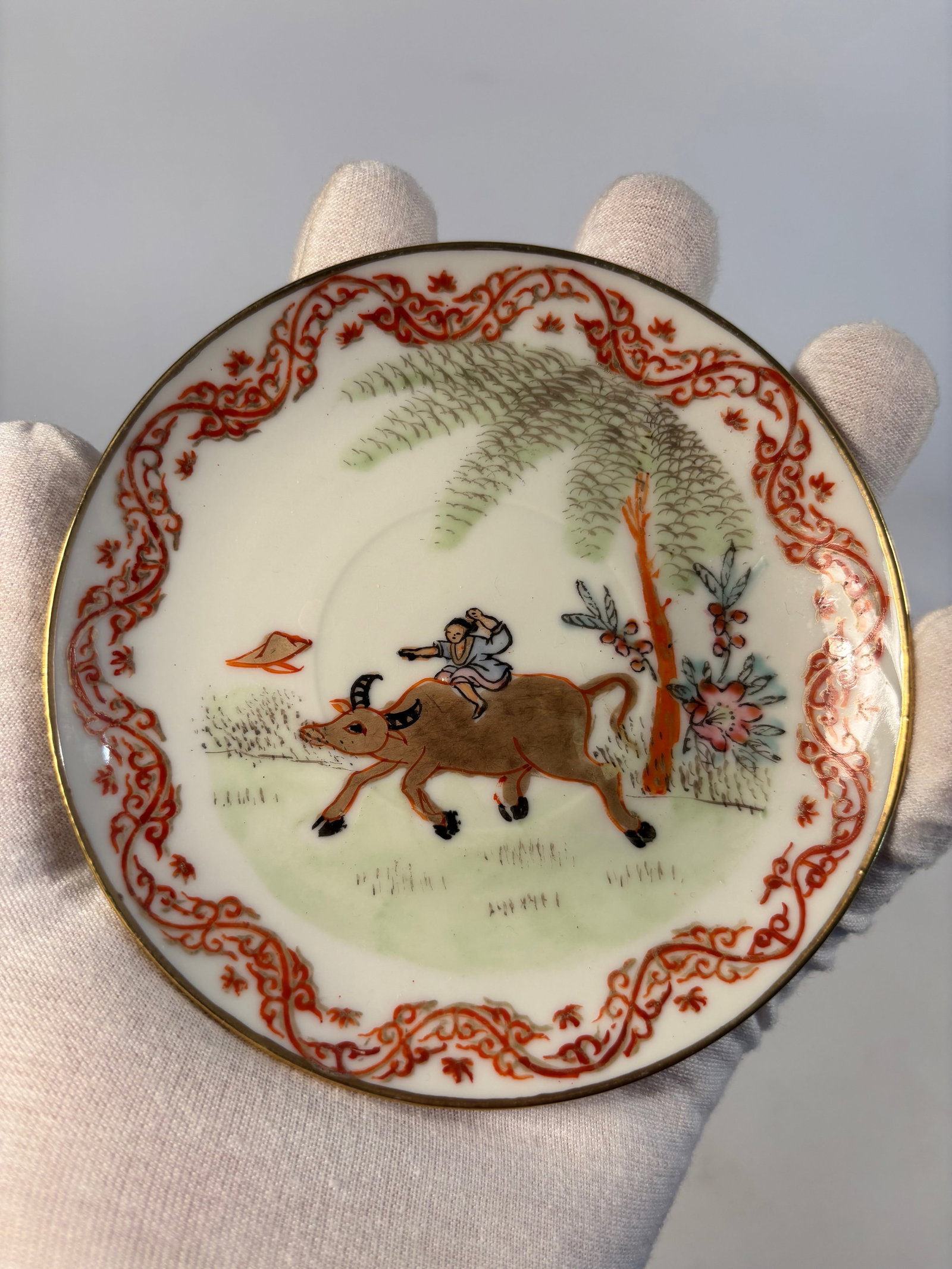 A Hand-Painted Porcelain Saucer with Boy on Water Buffalo: Late 19th century -early 20th century,The central scene depicts a boy riding a water buffalo beneath a large willow or palm tree, a classic Chinese pastoral motif symbolizing tranquility and harmony w