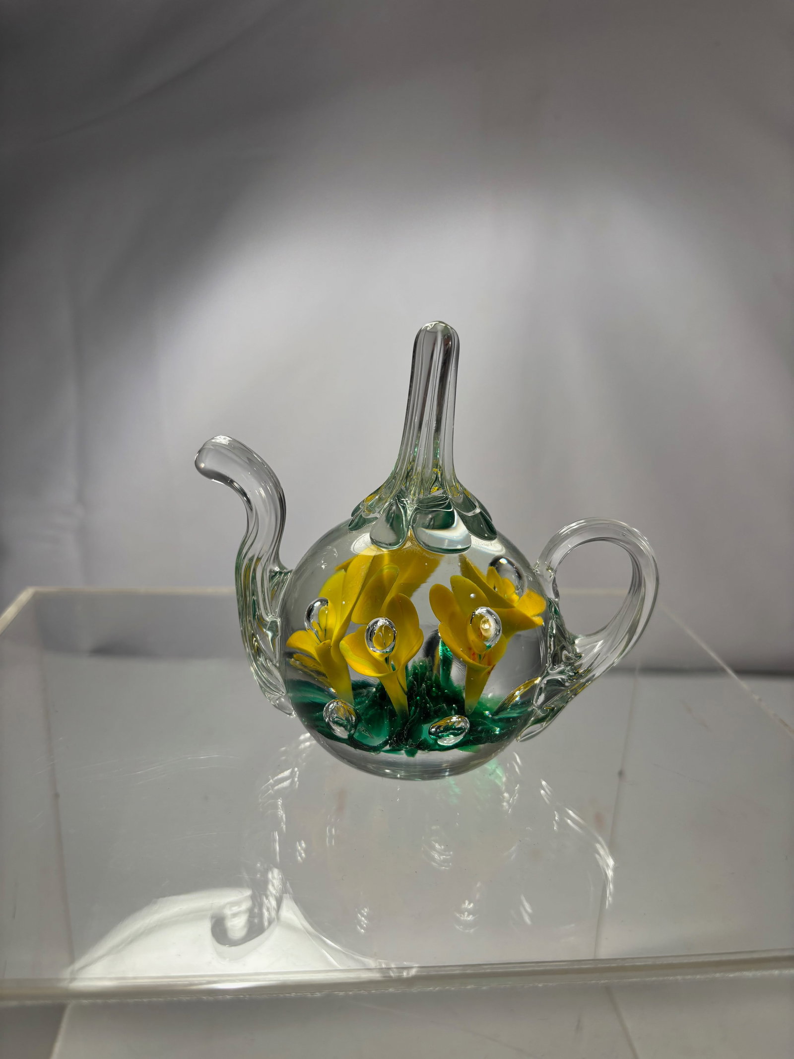 Studio Art Glass Miniature Teapot with Yellow Flowers: Studio art glass miniature teapot featuring yellow lampworked flowers with green foliage encased within clear glass, accented by controlled air bubbles. The piece is hand-formed with an applied spout,