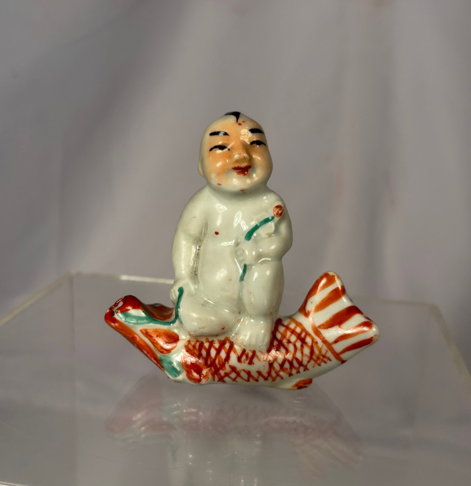 A Chinese Famille Rose Porcelain Figure of a Boy Riding a Carp: A Chinese famille rose porcelain figure depicting a smiling boy seated atop a carp, symbolizing prosperity, good fortune, and abundance in traditional Chinese culture. The figure is finely modeled and