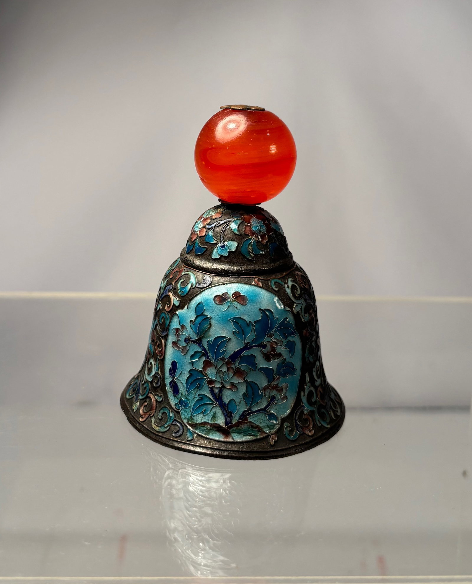 Chinese Cloisonné Enamel Bell with Red Glass Finial: 19th century, Chinese cloisonné enamel bell of flared form, decorated with scrolling floral and foliate motifs in shades of blue and turquoise against a dark enamel ground. The bell is surmounted by 