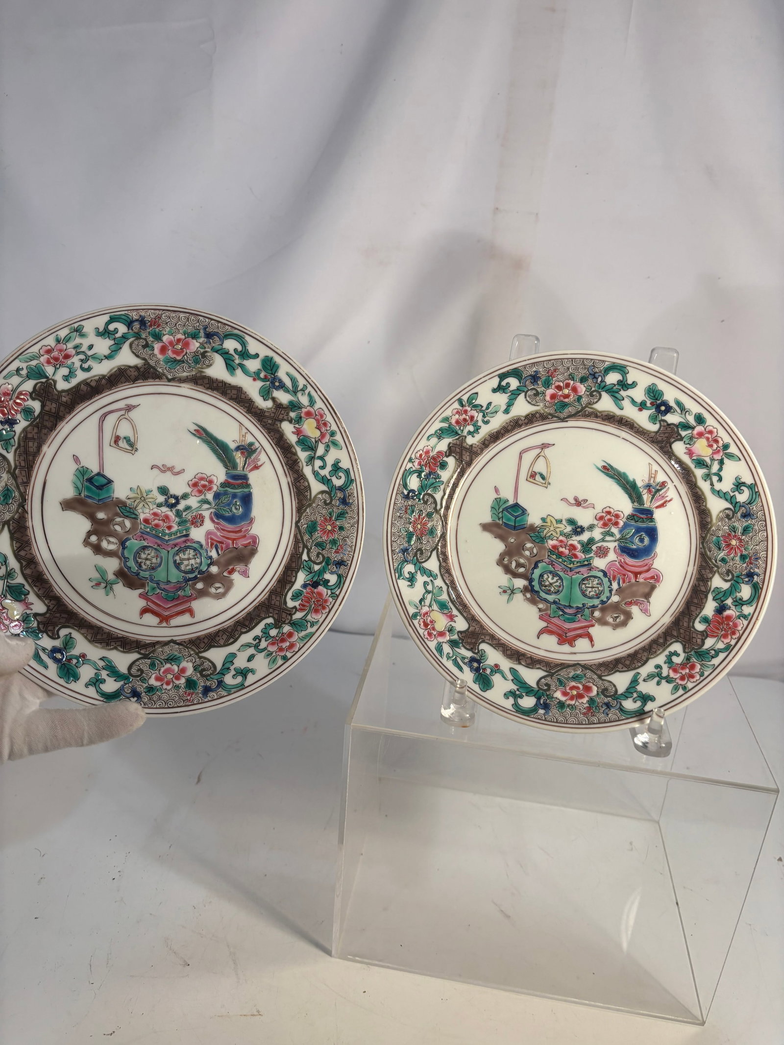 Pair of Japanese Polychrome Enameled Porcelain Plates with Precious Objects, Meiji: A pair of Japanese porcelain plates, each decorated in colorful enamels and gilt with an arrangement of vases, flowers, and auspicious precious objects. The rims feature dense floral-scroll and patter