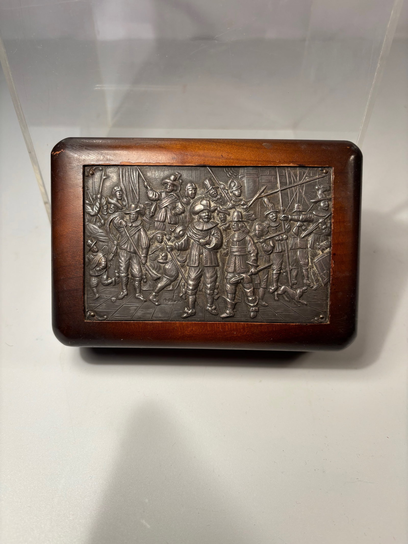 A Silver Relief Plaque Box Depicting Rembrandt’s “De Nachtwacht: A wooden box mounted with a silver relief plaque depicting a scene from The Night Watch by Rembrandt van Rijn. The plaque inscribed “De Nachtwacht” along the lower edge. The hinged wooden box 