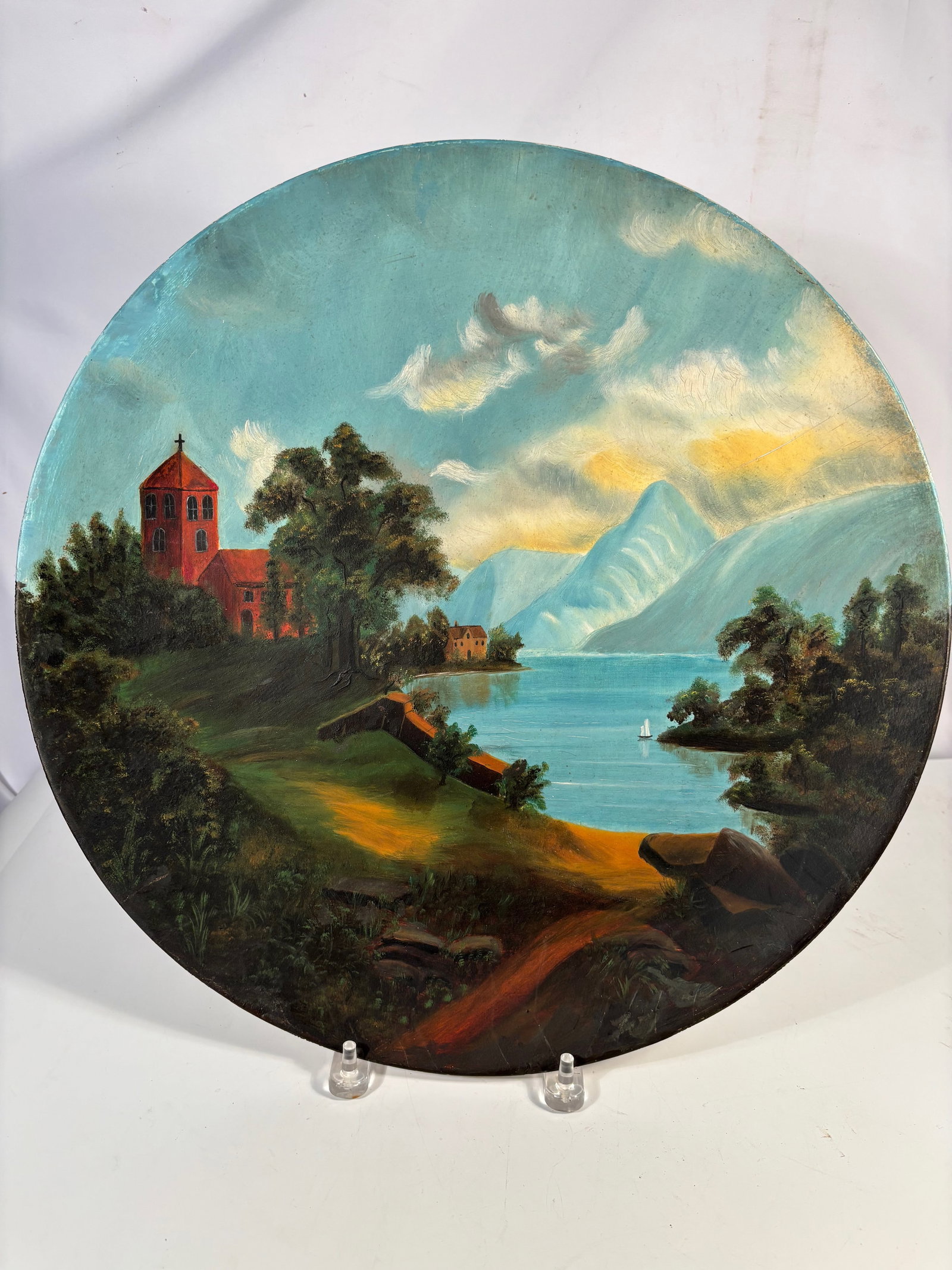 Vintage Large Round Canvas Painting of European Lake Landscape: Hand painted circular canvas depicting a European landscape with church, lake, mountains and sailing boat under a dramatic sky. Mounted on a round support panel and fitted with a hanging mount on the 