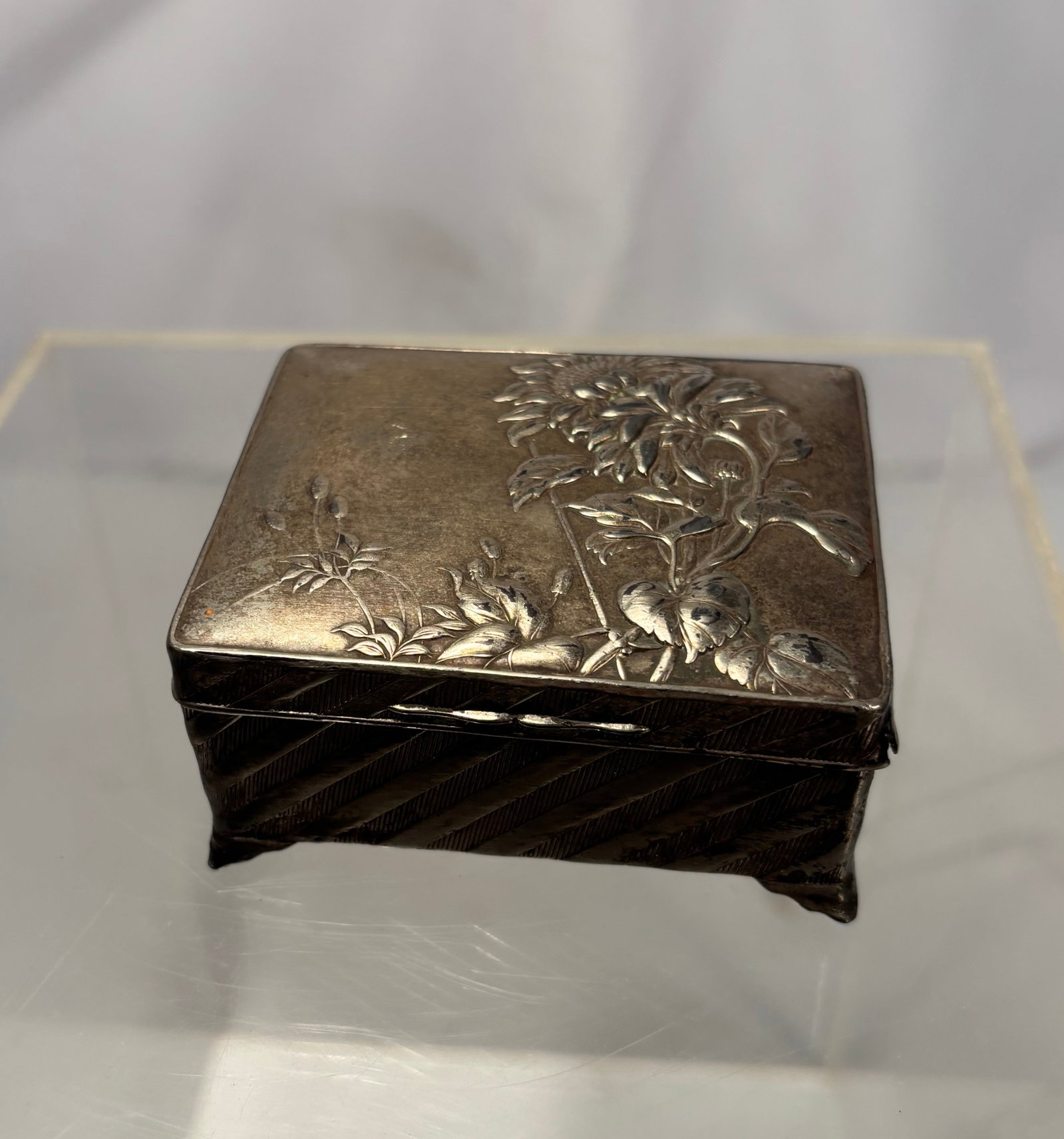 Antique Japanese Silvered-Metal Floral Trinket Box, Marked Japan Late Meiji to Taisho Period, circa: An antique Japanese export hinged box decorated on the lid with finely embossed and chased sunflower or chrysanthemum blossoms, foliage, and an insect motif. The sides feature engraved diagonal bands,