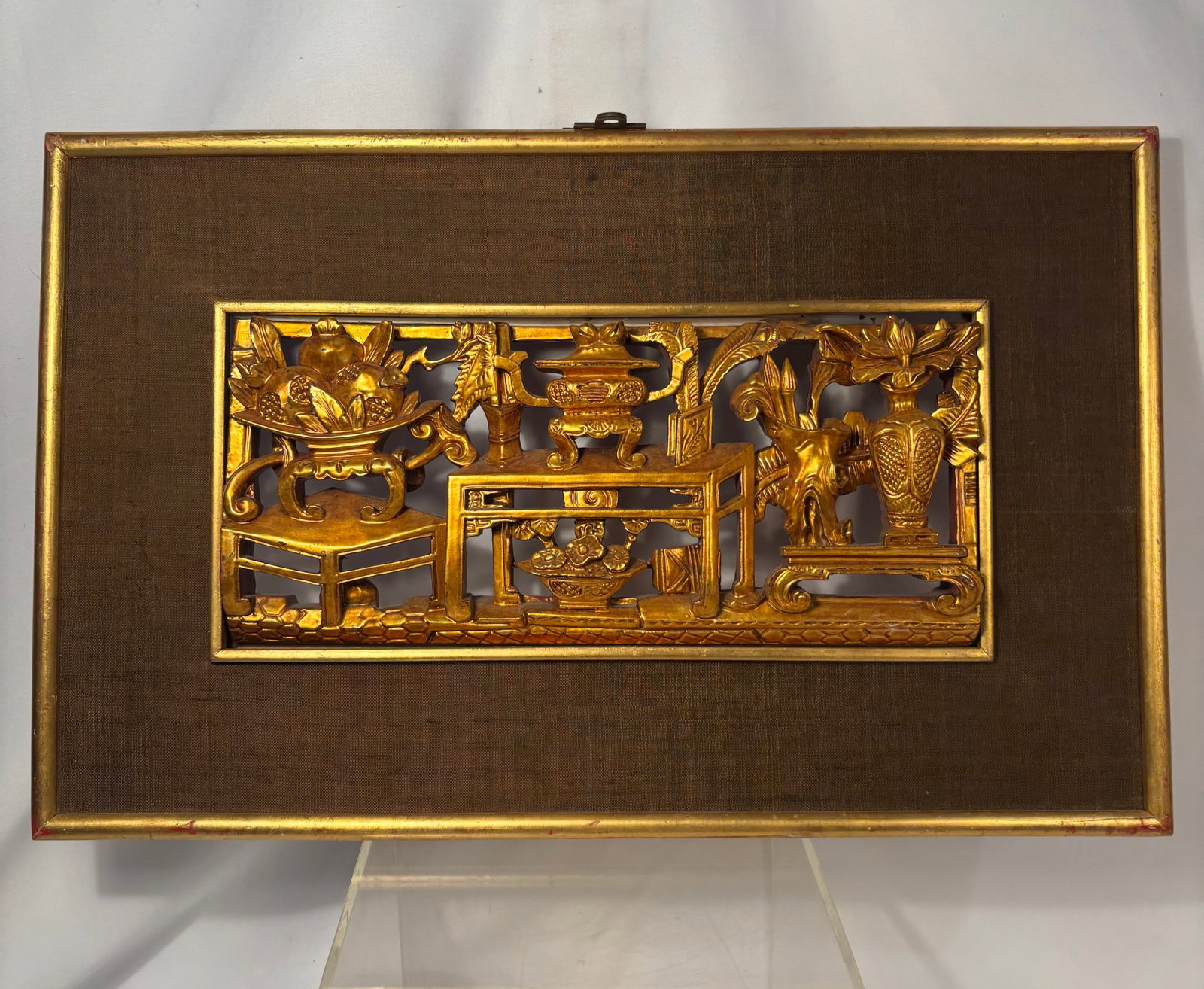 A Chinese Gilt-Carved Wood Scholar’s Objects Panel, Framed ,19th century: A Chinese carved and pierced wooden panel decorated with a traditional “Hundred Antiques” arrangement. The composition includes an incense burner, flower-filled vessels, fruit, scholar’s obj
