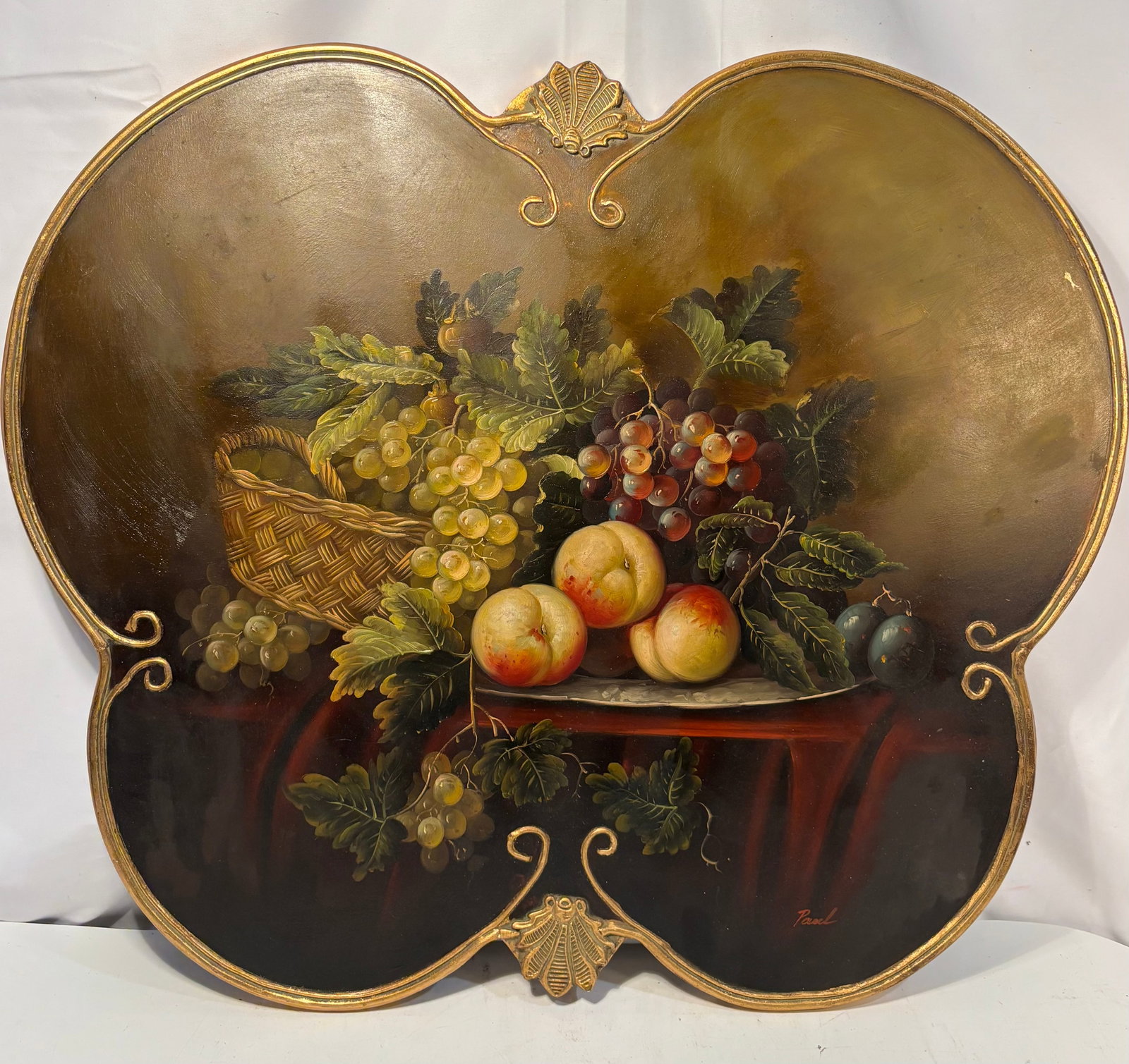 Vintage European-Style Fruit Still Life Oil Painting on Shaped Wood Panel: A decorative still-life painting depicting green and red grapes, apples or peaches, plums, foliage and a woven basket arranged upon a tabletop. Executed in oil on a shaped wooden panel with a metallic