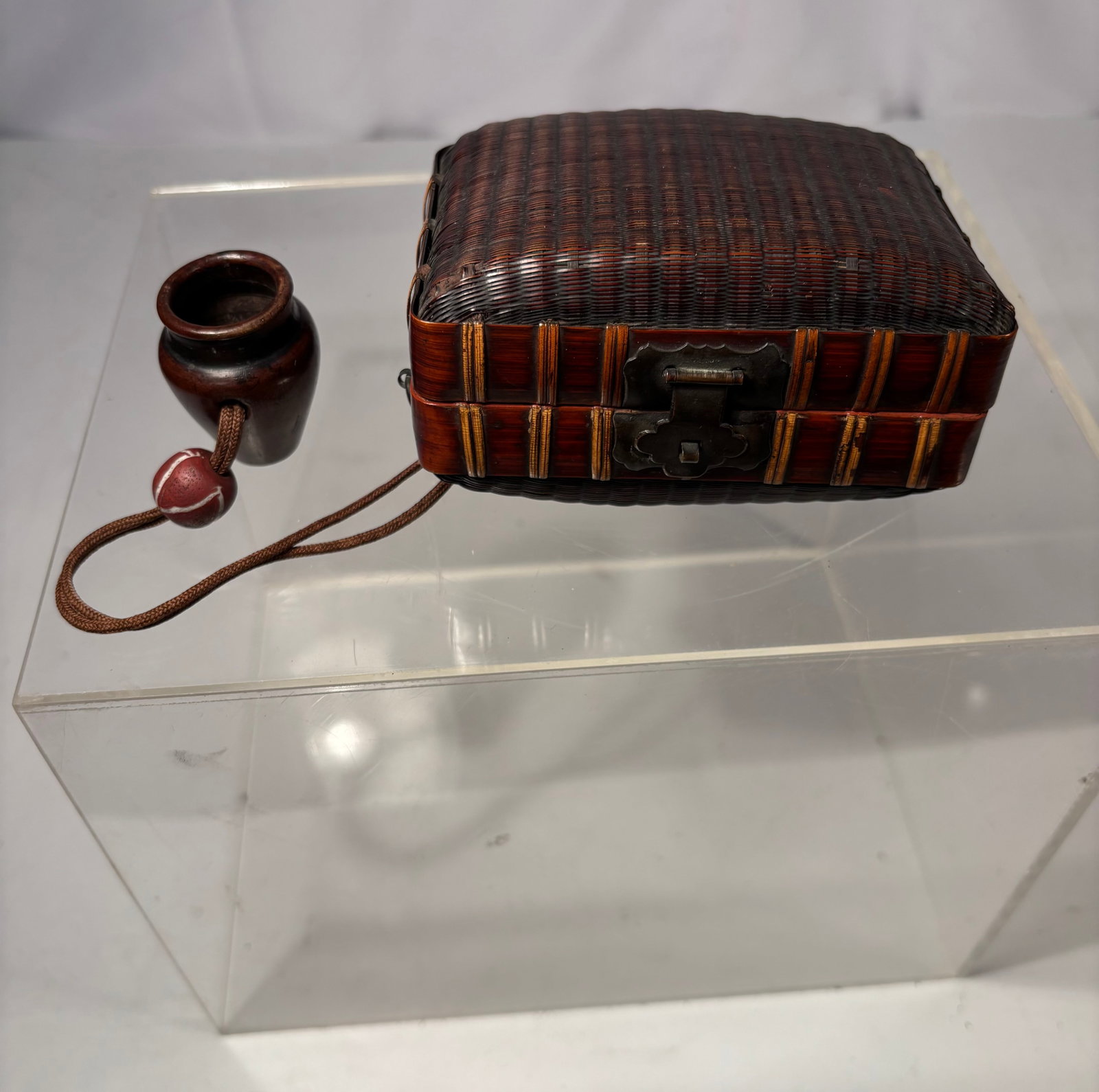 A Japanese Woven Bamboo and Lacquer Box with Cord and Pendant, Early 20th Century: A Japanese small box combining woven bamboo and lacquer elements, featuring a finely woven bamboo top with lacquered sides and decorative metal clasp. The piece retains its cord and pendant fittings a