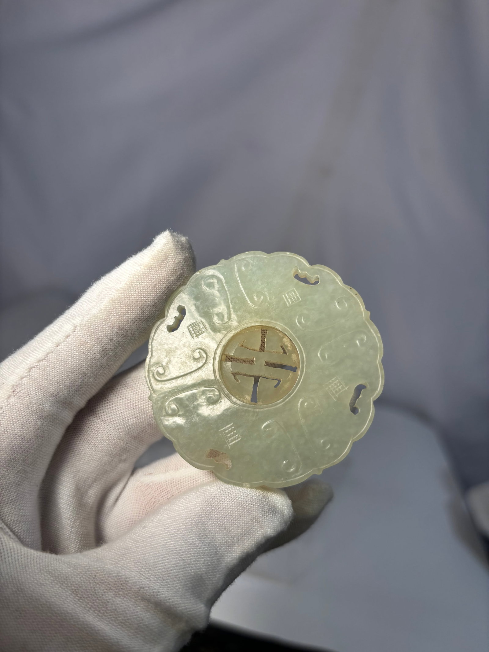 A Chinese Carved Celadon Jade Bi Disc with Shou Character: A finely carved Chinese jade bi disc, rendered in a pale celadon tone with semi-translucent quality.The circular plaque is centered with a carved “Shou” (longevity) character, surrounded by st