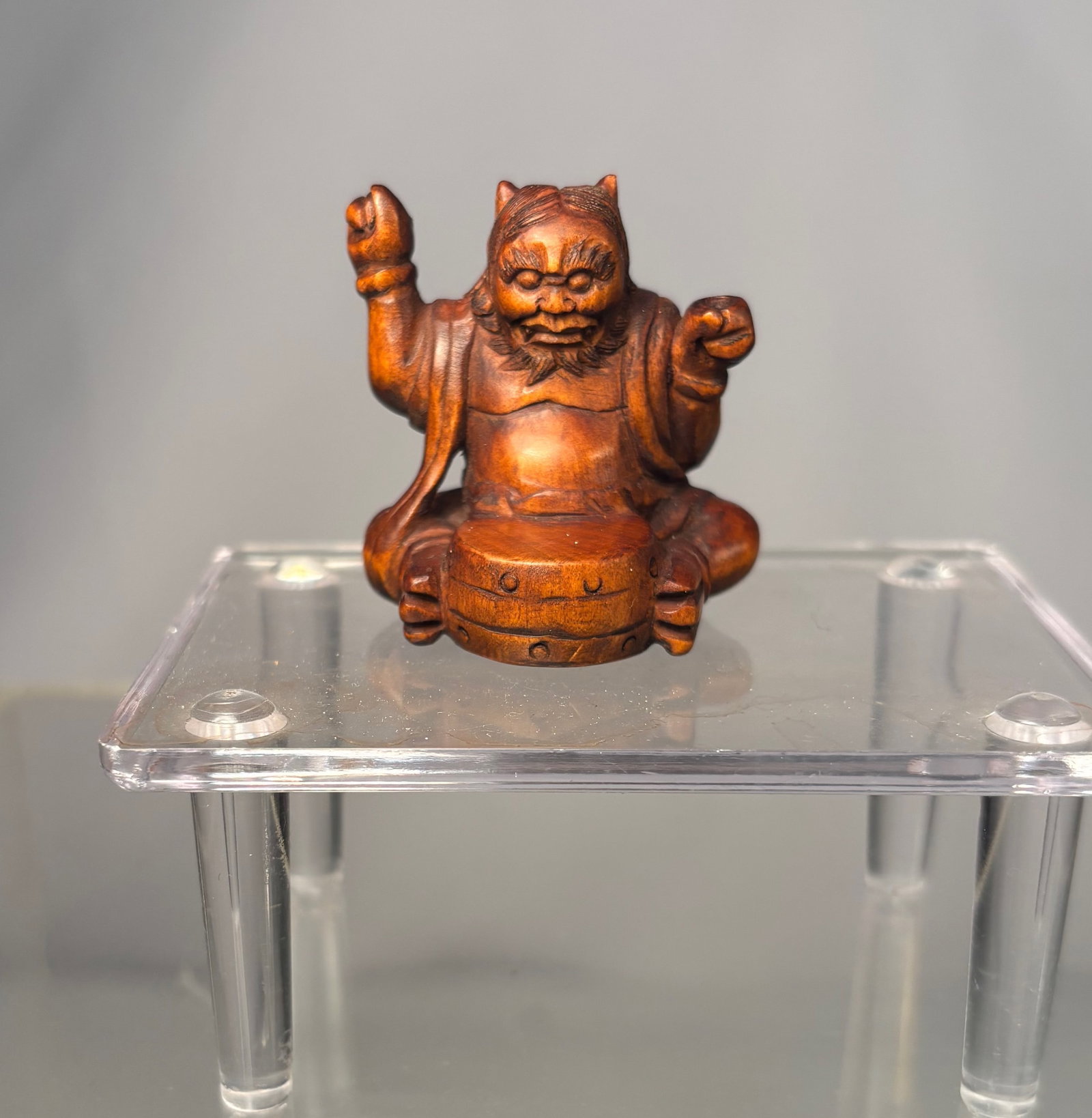 A Japanese Boxwood Netsuke of a Seated Oni on a Barrel, Signed (Gyokumatsu saku): Meiji period (19th century),A finely carved Japanese netsuke depicting a seated oni figure perched atop a barrel, shown with both arms raised in a lively and expressive pose. The figure is carved from