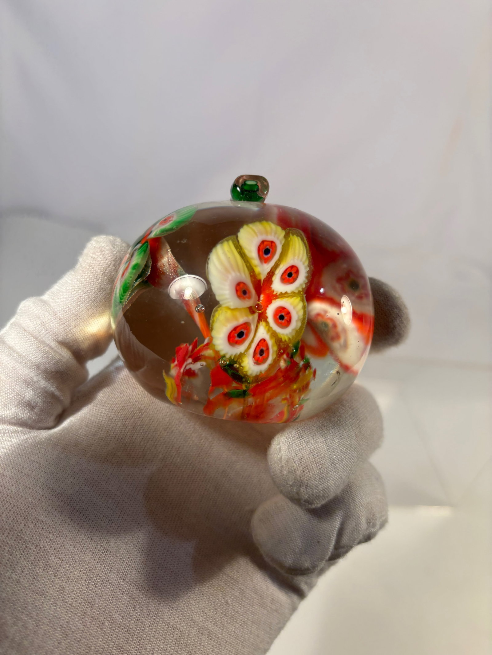A Murano Style Millefiori Art Glass Apple Paperweight with Applied Stem: Decorative art glass paperweight formed as an apple, featuring colorful millefiori floral canes in red, green, and white tones suspended in clear glass. Finished with an applied green glass stem. Diam