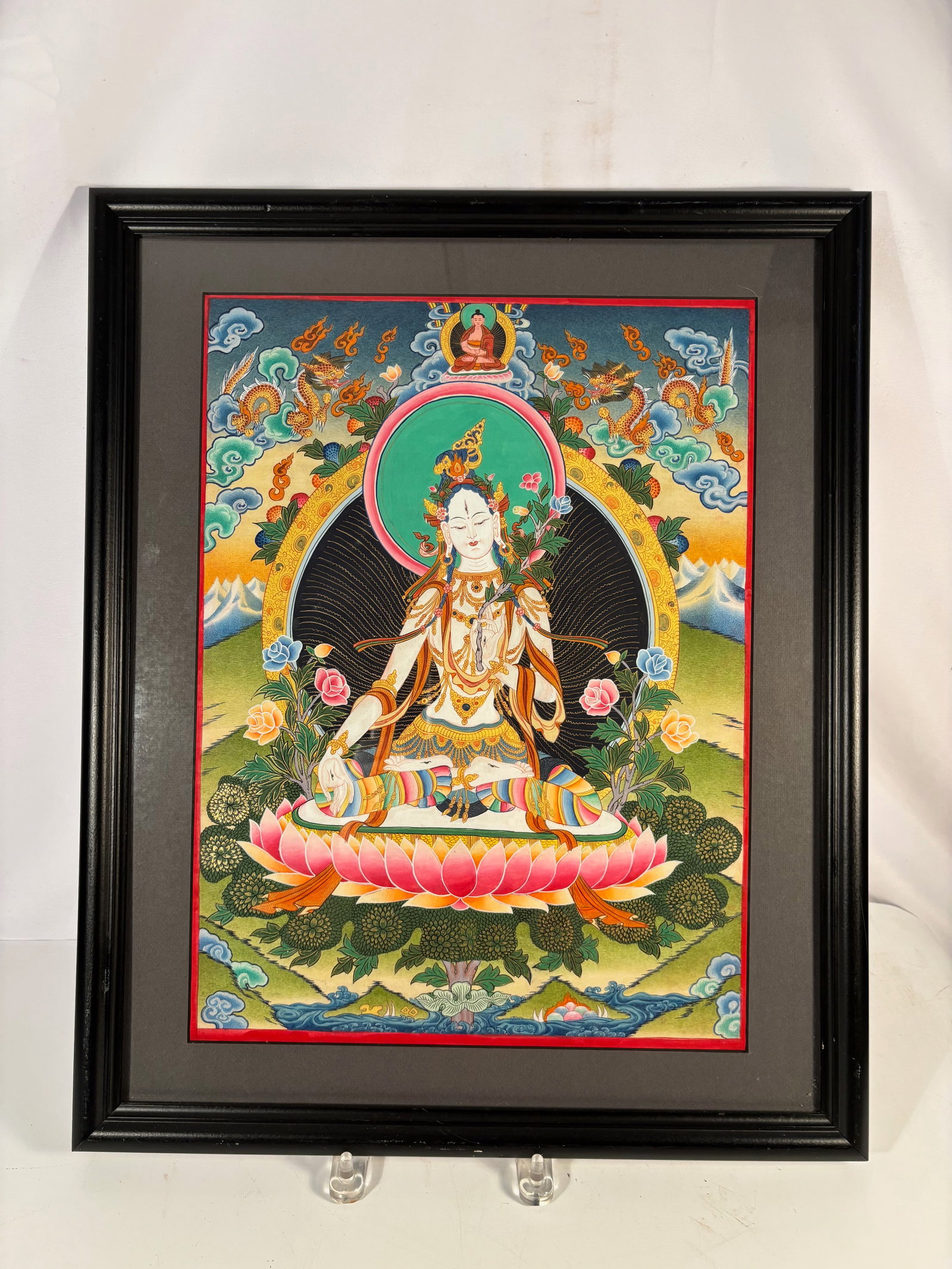 A Tibetan Style White Tara Thangka: Tibetan style thangka depicting White Tara seated on a lotus throne, surrounded by dragons, clouds, and floral motifs. Mounted and framed. Frame Dimension: 18 x 22 inches.