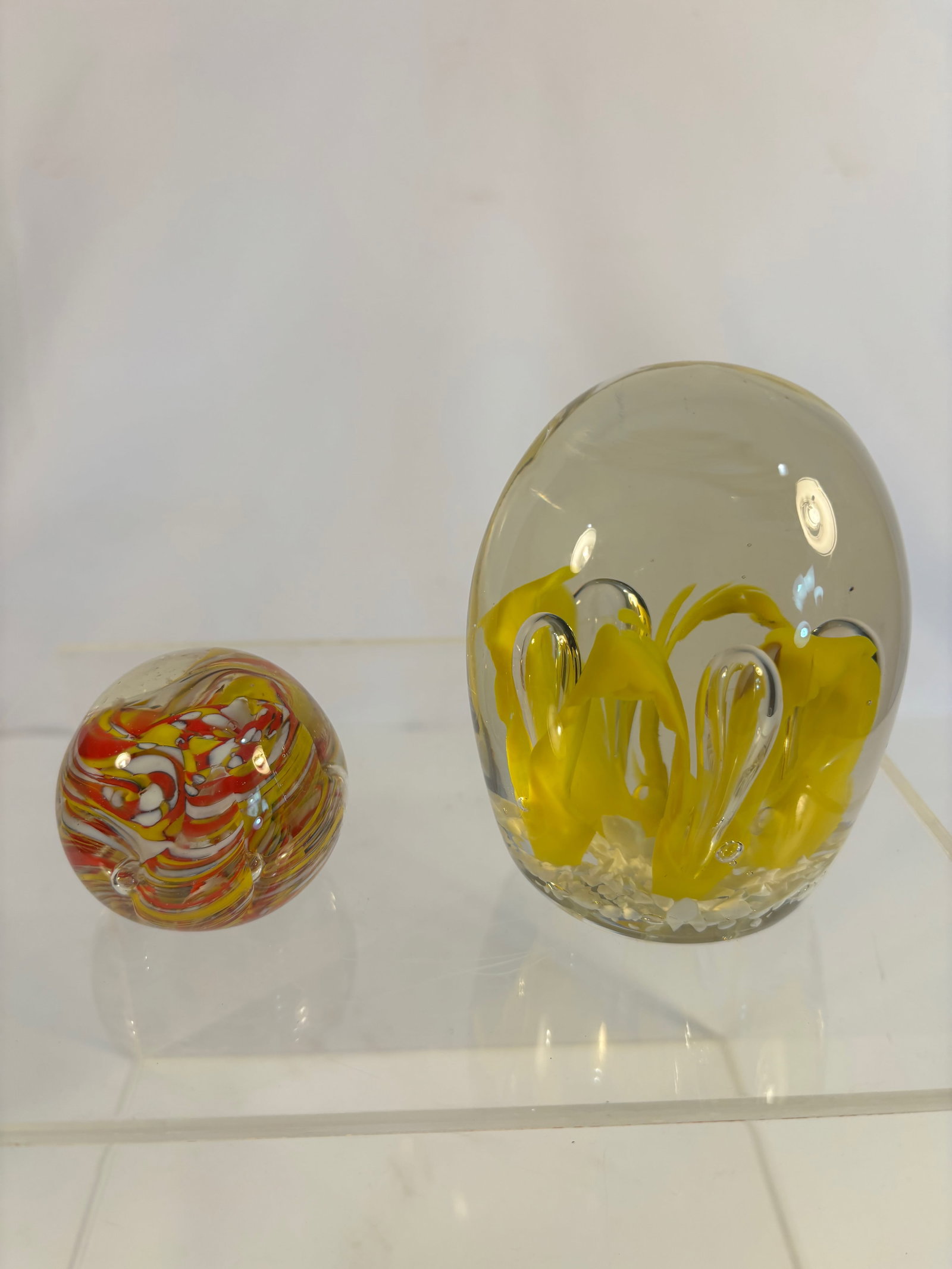 Lot of 2 Studio Art Glass Paperweights — Floral Designs, Including Yellow and Pink Lampwork: Lot of 2 studio art glass paperweights, each showcasing delicate lampwork floral designs encased in clear crystal domes.The first features a soft pink blossom with green leaves rising from the base, f