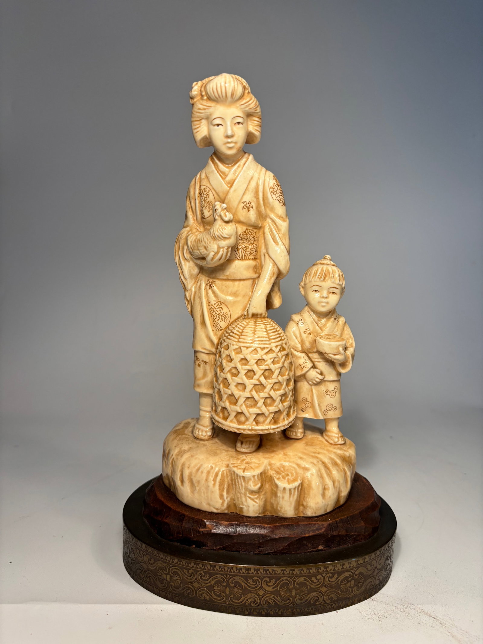 A Japanese Moriyama porcelain statue of woman and child: LATE 19TH, EARLY 20TH Century; Depicting a standing woman in kimono holding a rooster, accompanied by a small child holding a bowl. A large woven-style basket sits between them on a rocky base, all mo