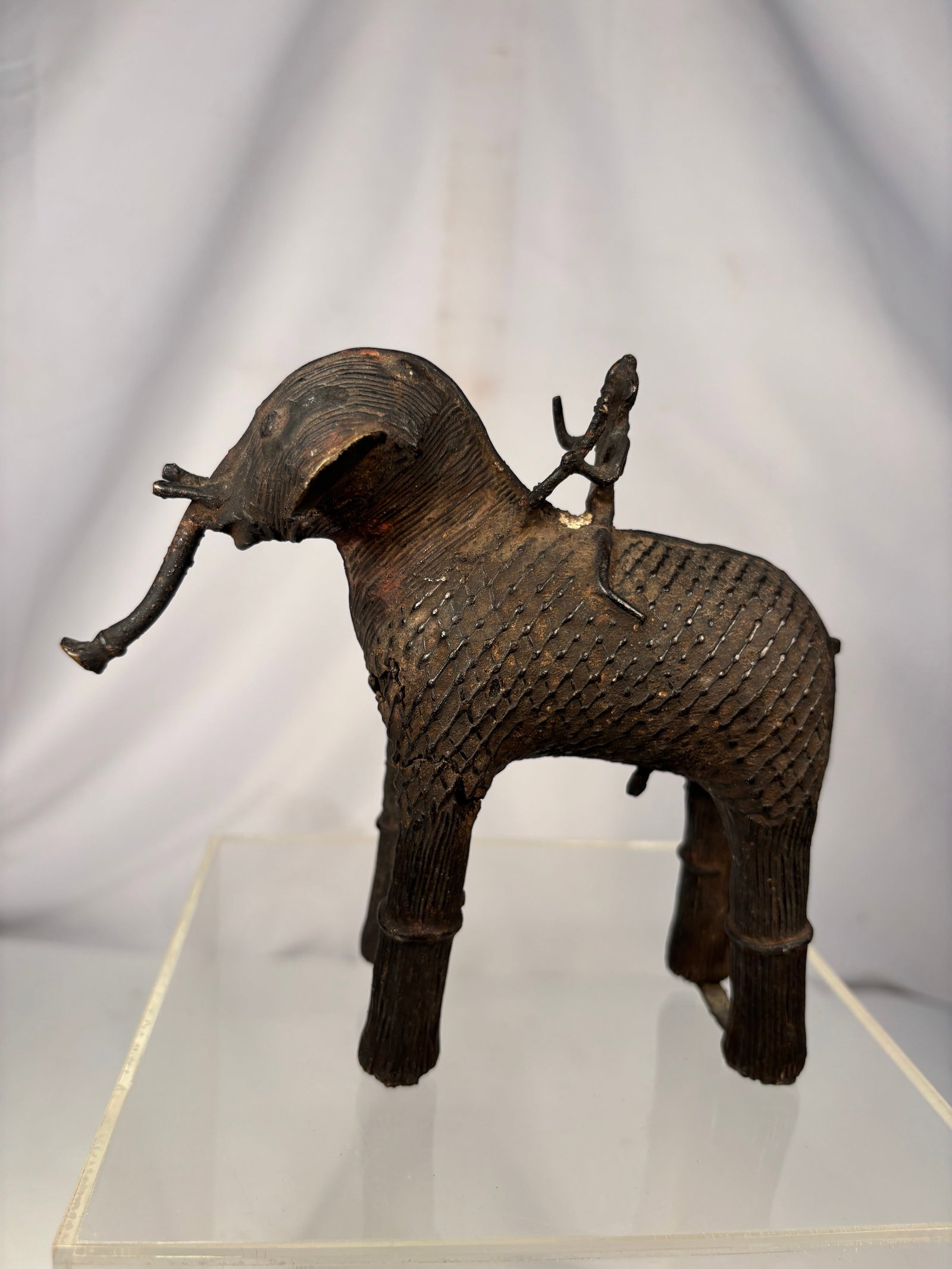 West African Bronze Elephant Rider Figure: Hand-cast African bronze sculpture depicting an elephant with a rider seated on its back. Created using the traditional lost-wax casting method, the figure features incised geometric decoration, a rai
