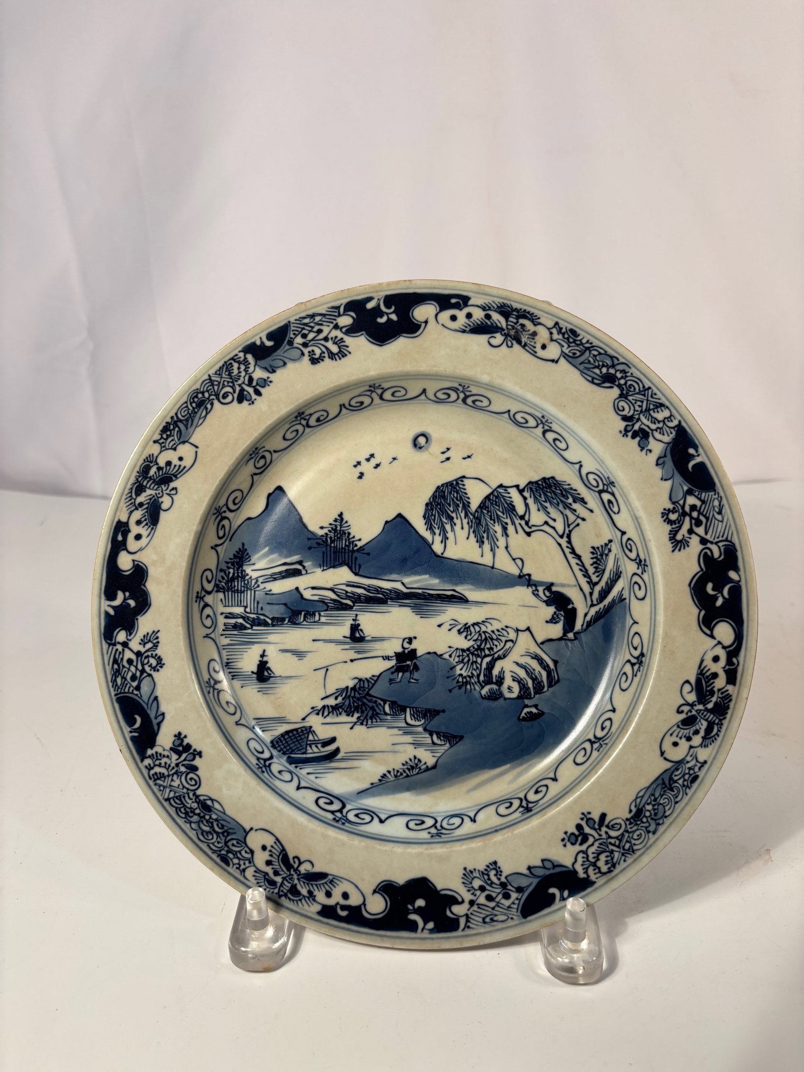 A Japanese Blue-and-White Porcelain Plate Meiji Period (c.1880–1910), Square Seal Mark: Finely painted Japanese blue-and-white porcelain plate depicting a river landscape with figures, pavilions, and sailing boats within a scrolling floral border. Glazed in soft white with deep cobalt to