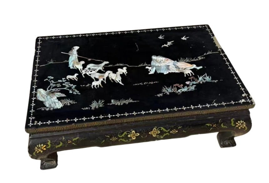 An Asian Black Lacquer Low Table with Mother-of-Pearl Inlay and Red-Lacquered Base: Early 20th century, Asian black lacquer low table, the rectangular top inlaid with mother-of-pearl depicting a pastoral scene of figures, animals, and birds within a geometric key-fret border. The apr