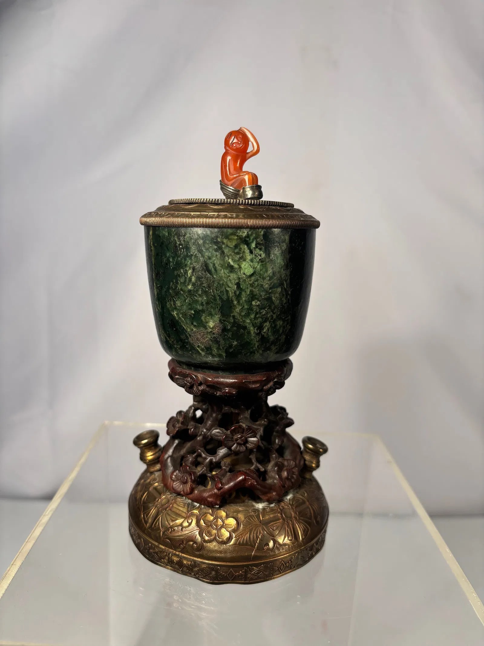 A Chinese Spinach Jade Inkwell with Gilt Bronze Stand and Cornelian Agate Monkey Finial: 19th century; A Chinese spinach jade inkwell of cup form, carved from dark green stone with natural mottling. The vessel is fitted with a gilt bronze cover surmounted by a finely carved cornelian agat