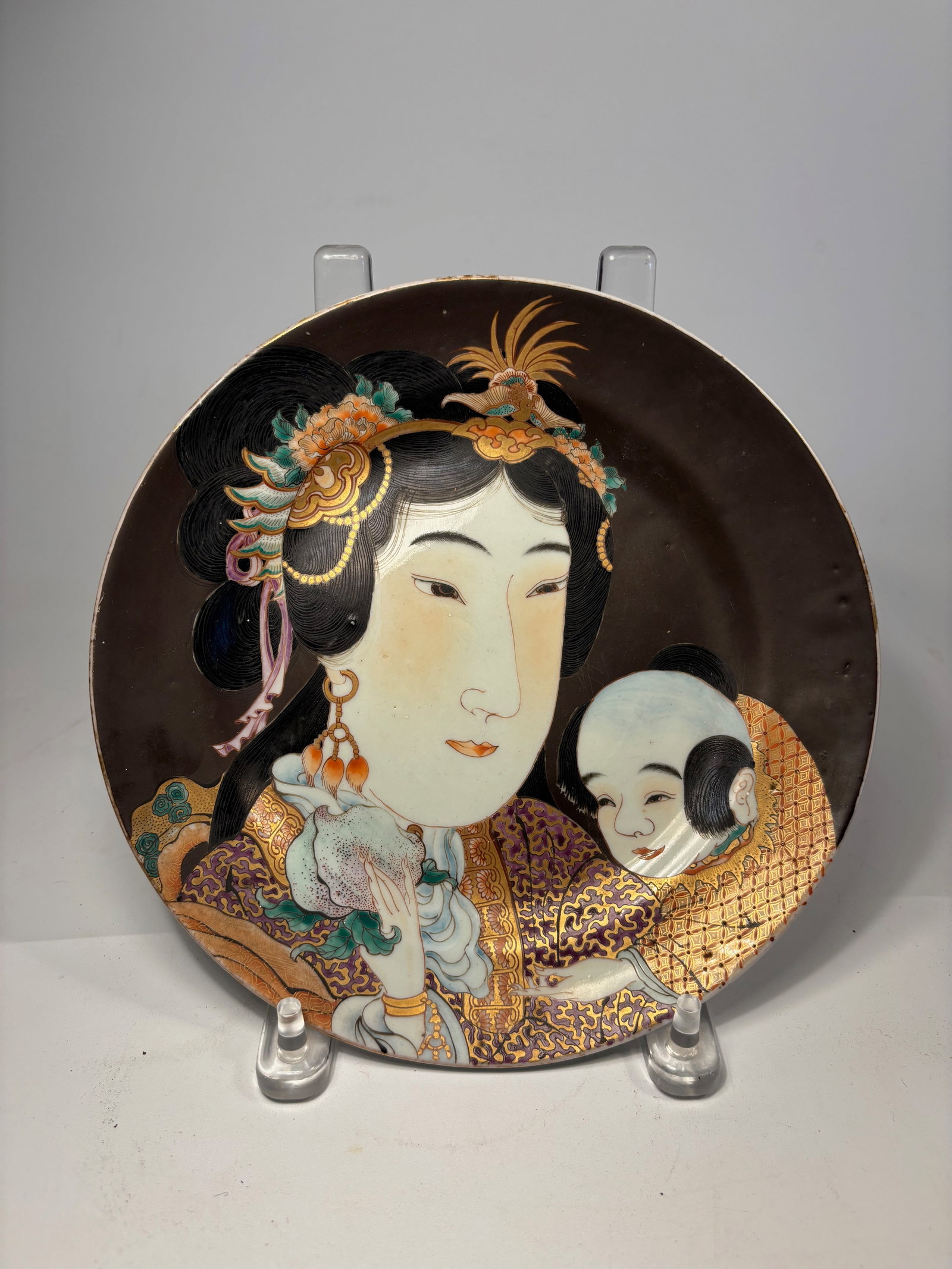 A Japanese Satsuma-Style Porcelain Plate – Bijin with Child, Hand-Painted & Gilt: Meiji-Period(1868-1912); The design features an elegant bijin (beautiful woman) in elaborate courtly attire, adorned with ornate hair ornaments, holding a delicate fan. Beside her is a smiling child w