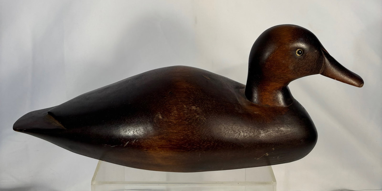 A vintage Hand-Carved Wooden Duck Decoy with Glass Eyes: Featuring a finely shaped head and body with inset glass eyes. The decoy displays a rich, dark patina developed through age and use, with visible hand-tool marks and natural asymmetry consistent with 