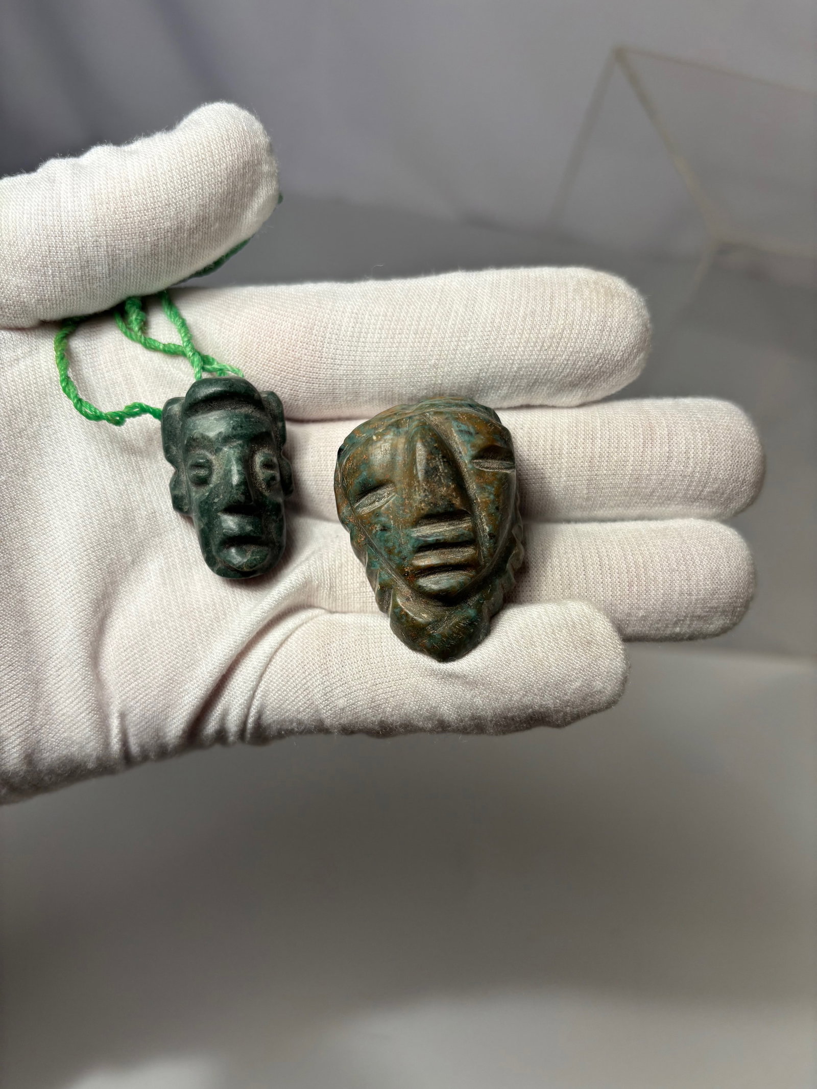 Two Pre-Columbian Jade/Greenstone Miniature Masks, Olmec to Maya Style: Each carved from mottled greenstone, depicting stylized human faces with pierced tops for suspension, likely worn as amulets or pendants. One of elongated form with high relief features and squared ba
