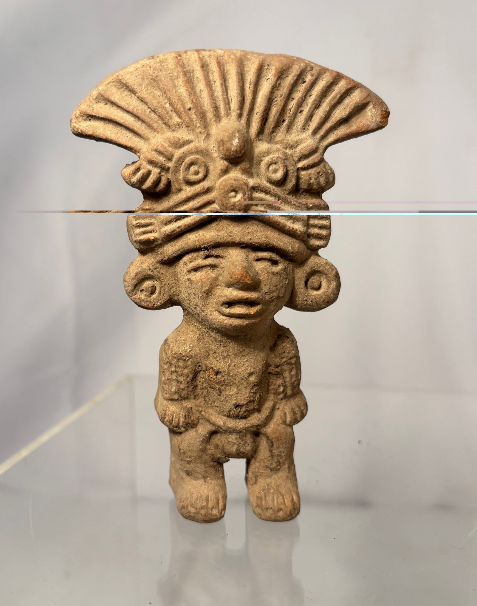 Pre-Columbian Terracotta Standing Figure: Terracotta standing figure in the style of ancient Pre-Columbian Mesoamerican art, featuring a figure with an elaborate fan-shaped headdress and incised details. Height: 5 in. .