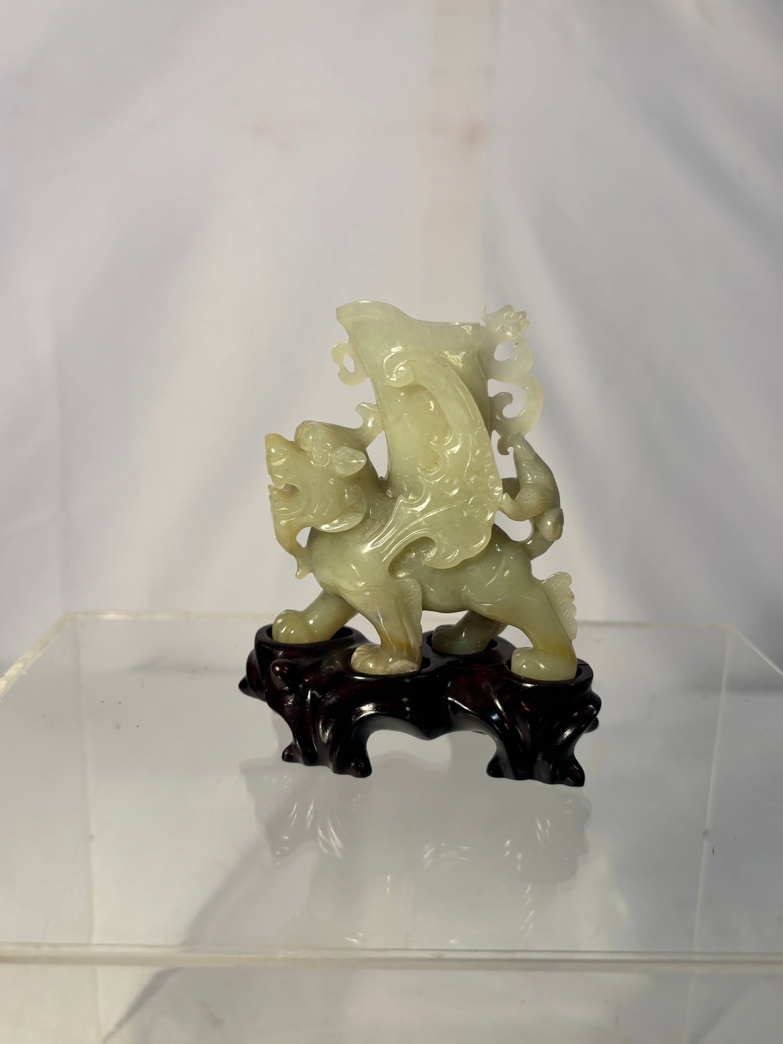 A Chinese Carved Jade Figure of Mythical Beast with Vase, on Wood Stand: Qing Dynasty(1644-1912) ; A Chinese carved jade stone figure depicting a mythical beast (possibly a pixiu) supporting a vase form, symbolizing prosperity and protection. The carving is rendered in a p