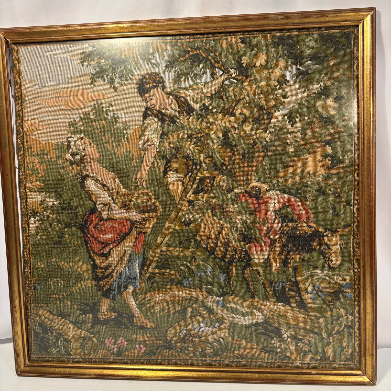 Vintage Framed Gobelin-Style Woven Tapestry Depicting a Pastoral Fruit-Gathering Scene Early to mid: A vintage European-style woven tapestry, often referred to as a gobelino, depicting a romanticized pastoral scene with a rural couple gathering fruit beneath a tree, accompanied by a laden donkey. Ren