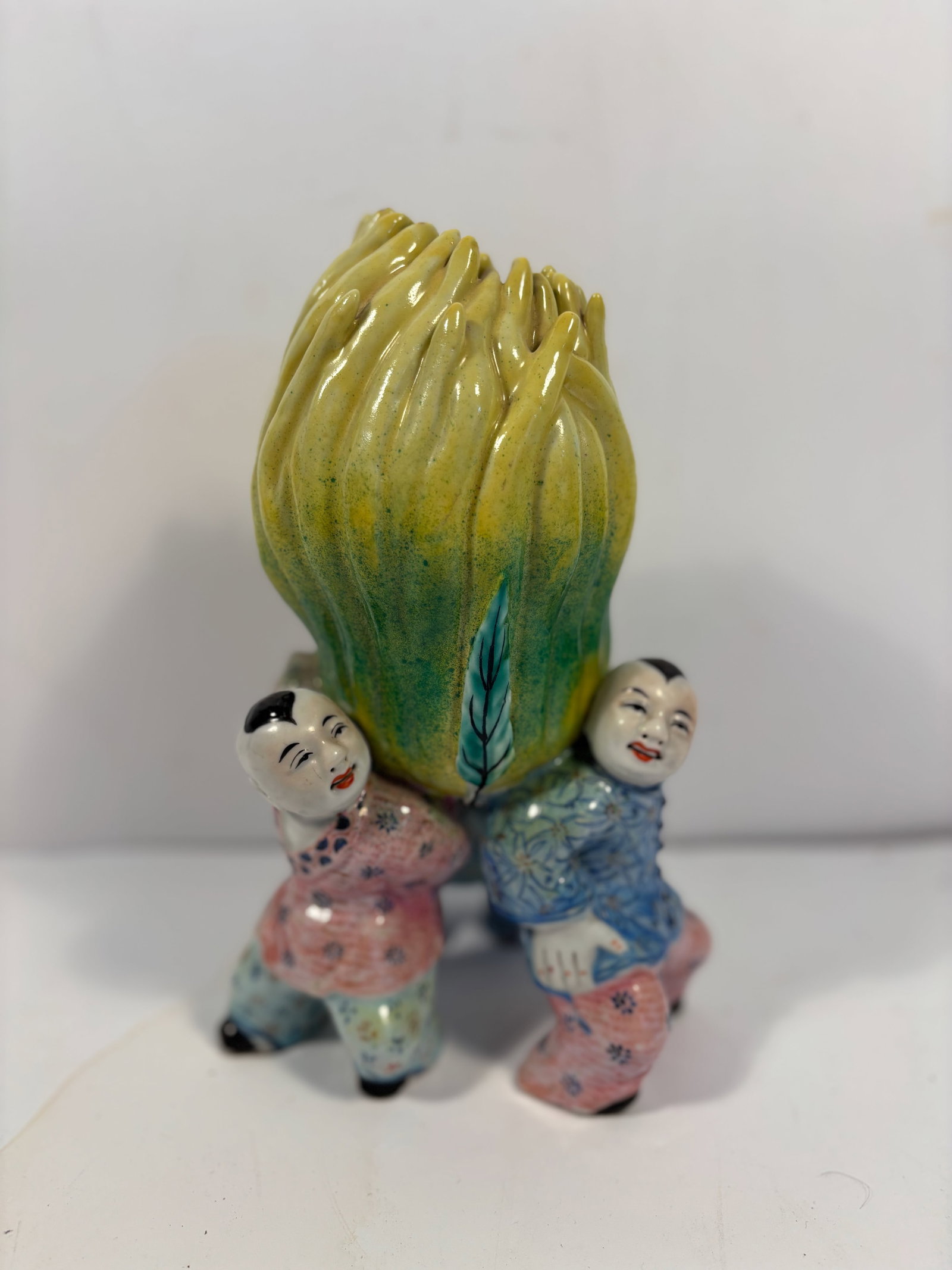 A Chinese Famille Rose Porcelain Figurine: 19th century; 19th century; A beautifully crafted Famille Rose porcelain figurine depicting three boys joyfully holding a Buddha's Hand Dimensions?HEIGHT: 8 inches; Width: 6 inches; Depth: 5 inches.