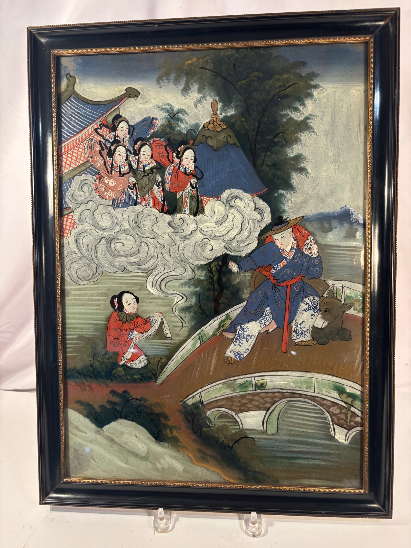 Antique Chinese Reverse-Painted Glass Figural Scene, Framed, possibly 19th century: A Chinese reverse painting on glass depicting a colorful mythological or theatrical scene. A male figure stands upon an arched bridge accompanied by a recumbent ox, while a female attendant kneels nea