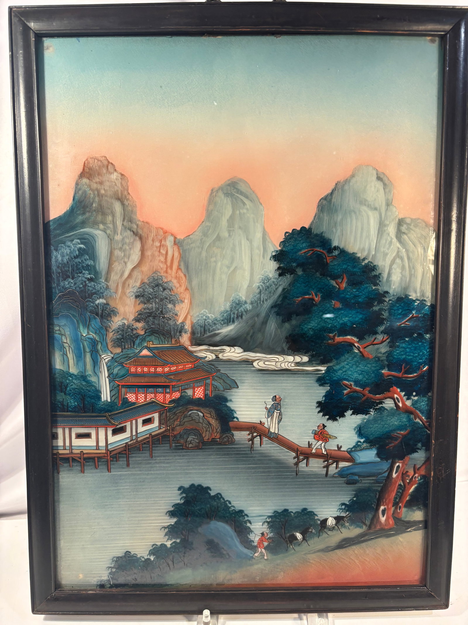 Antique Chinese Reverse-Painted Glass Landscape, Framed , possibly 19th century: A Chinese reverse painting on glass depicting an expansive mountainous river landscape. The scene features towering peaks, a waterside pavilion, a footbridge with two figures, cascading waterfalls, an