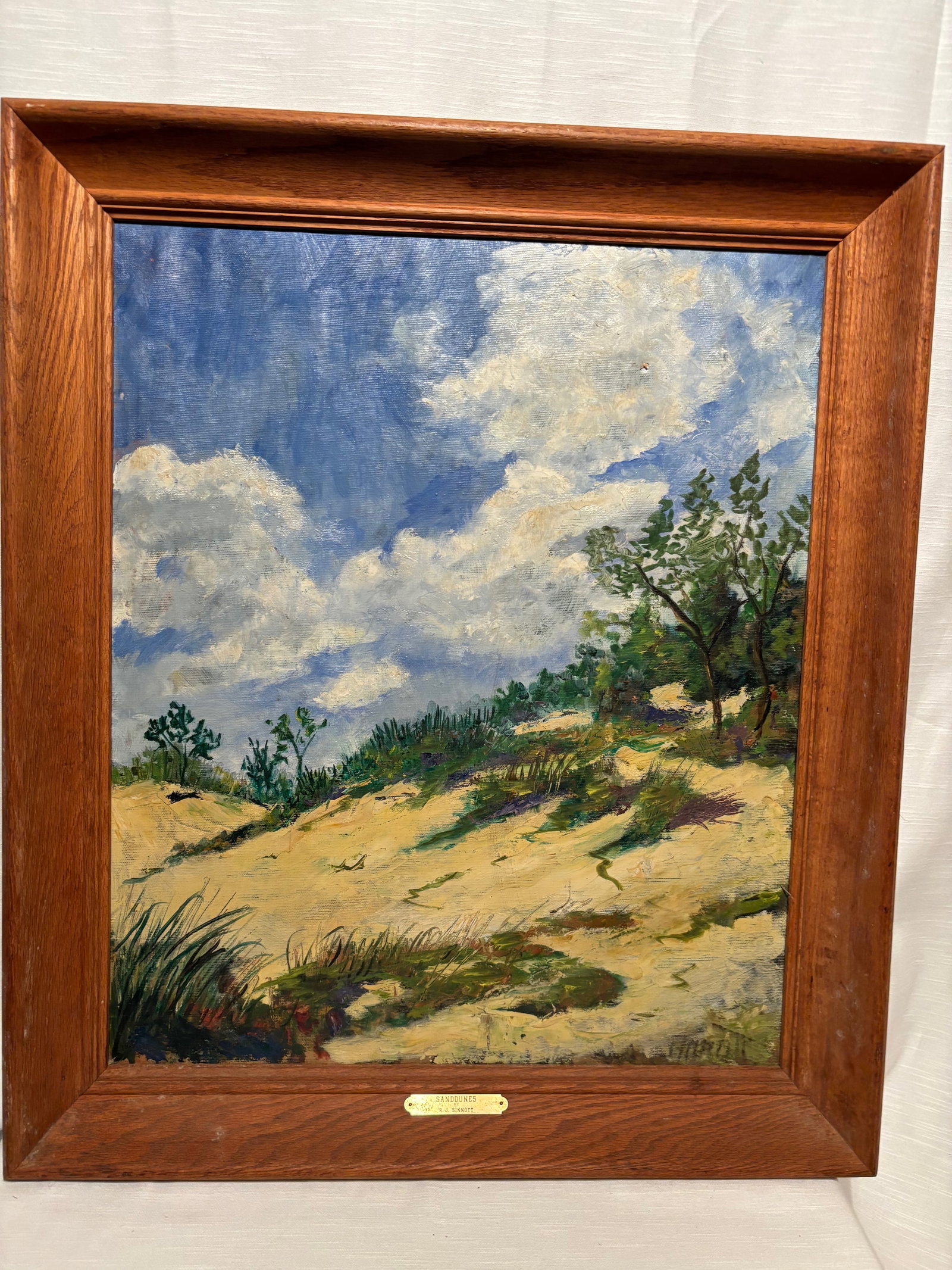 Vintage Coastal Sand Dunes Oil Painting, Signed R.J. Sinnott, Framed Beach Landscape: A vintage oil painting titled “Sand Dunes” signed R.J. Sinnott, depicting a tranquil coastal landscape with rolling dunes, windswept grasses, and an expansive sky filled with soft clouds. The 