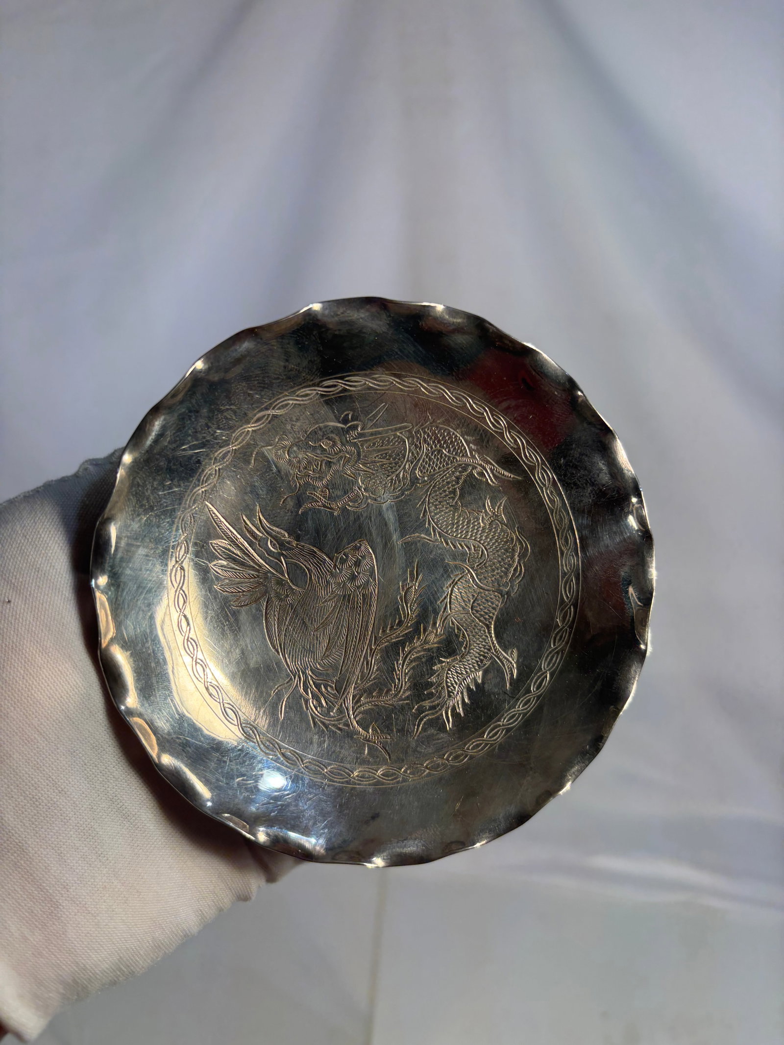 Sterling Silver Dragon and Phoenix Footed Dish, 20th Century: A sterling silver footed dish of scalloped circular form, raised on three applied feet. The interior is finely engraved with a traditional dragon and phoenix motif within a decorative border, symboliz