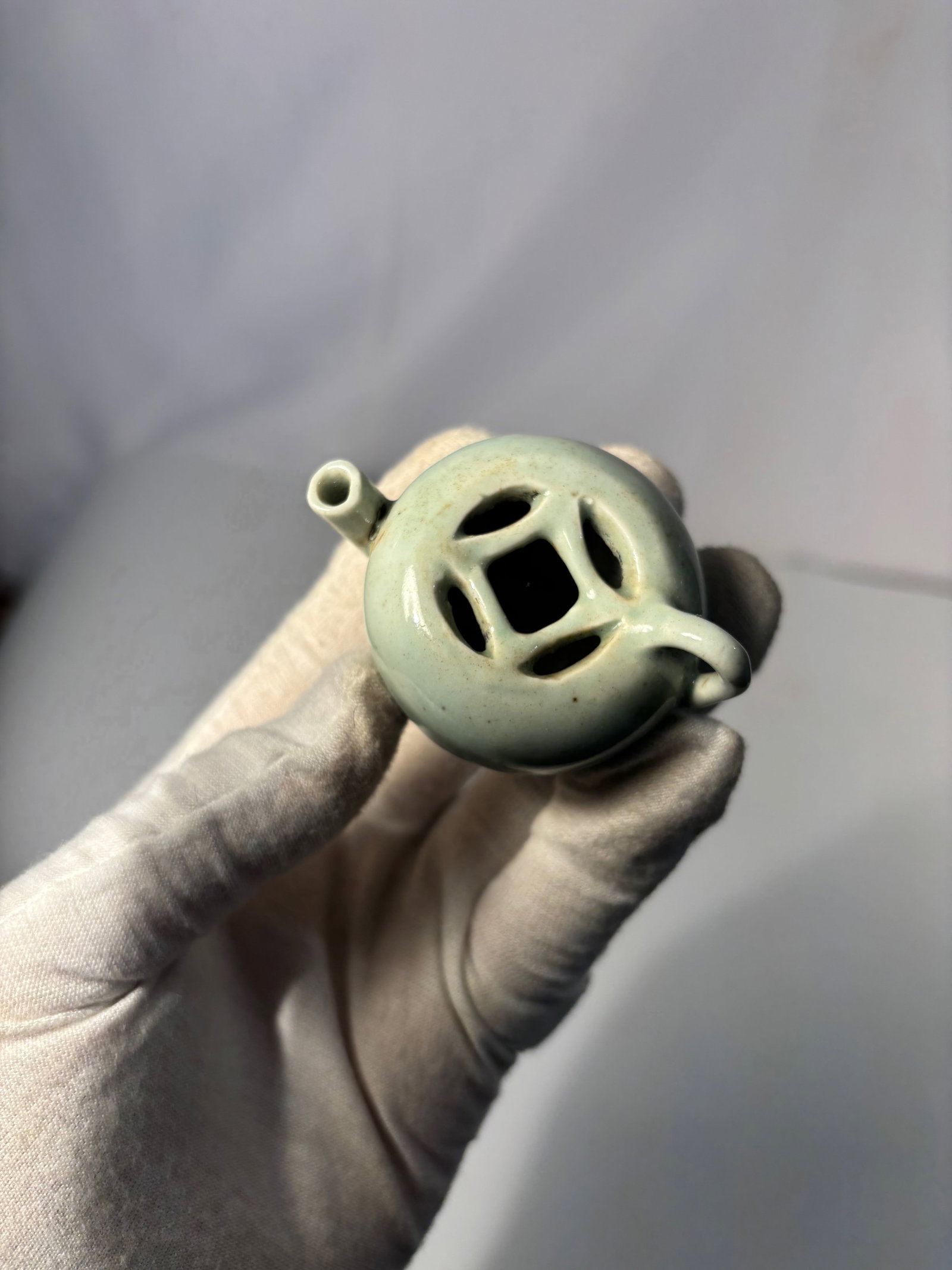 A Chinese Celadon-Glazed Miniature Water Dropper, Probably Ming Dynasty: A finely potted Chinese miniature celadon-glazed vessel, likely intended as a scholar’s object, such as a small incense burner or water dropper. The vessel is formed with a compact, rounded body and