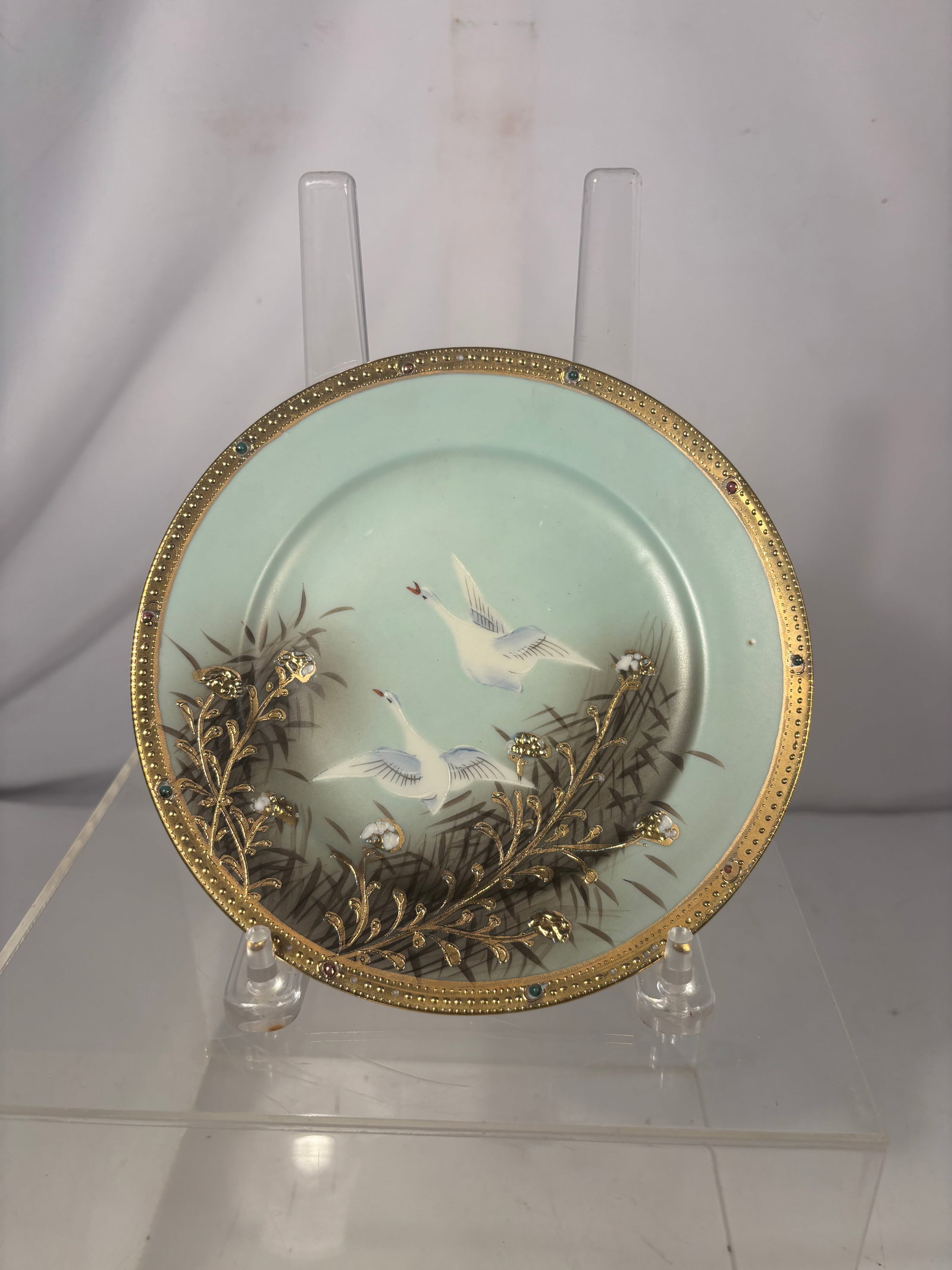 Japanese Hand-Painted Porcelain Plate with Cranes, Gilt-Metal Overlay and Jeweled Rim: A Japanese porcelain decorative plate depicting two cranes flying above bamboo and flowering plants on a pale celadon-toned ground. Embellished with applied gilt-metal foliate decoration, floral eleme