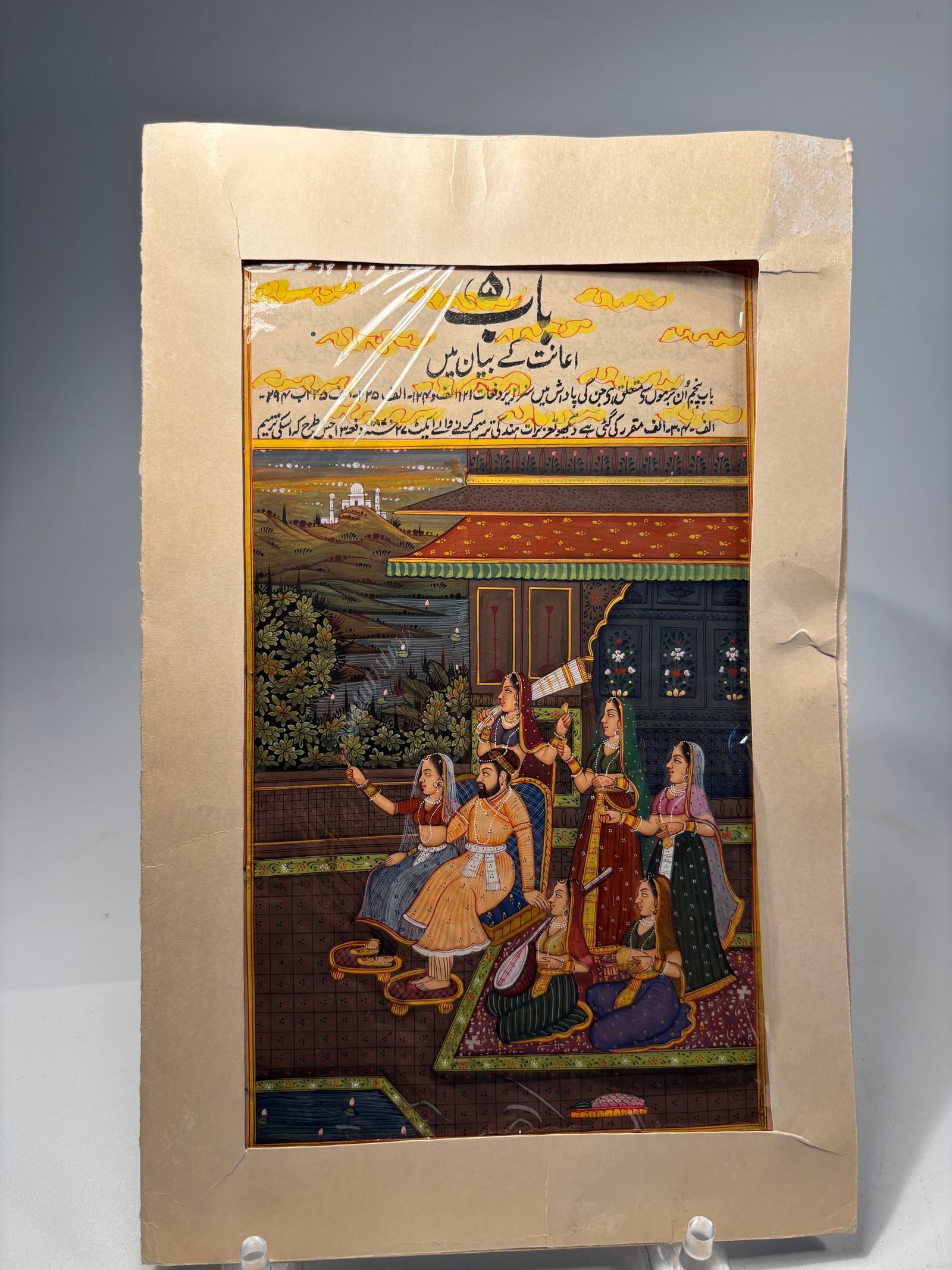 A Mughal Royal Court Miniature with Courtesans and Musician, Urdu Inscription, India: 19th century; A finely detailed Mughal-style miniature painting, gouache on paper, featuring a richly dressed noble seated within a palace terrace surrounded by courtesans and attendants. One attendan
