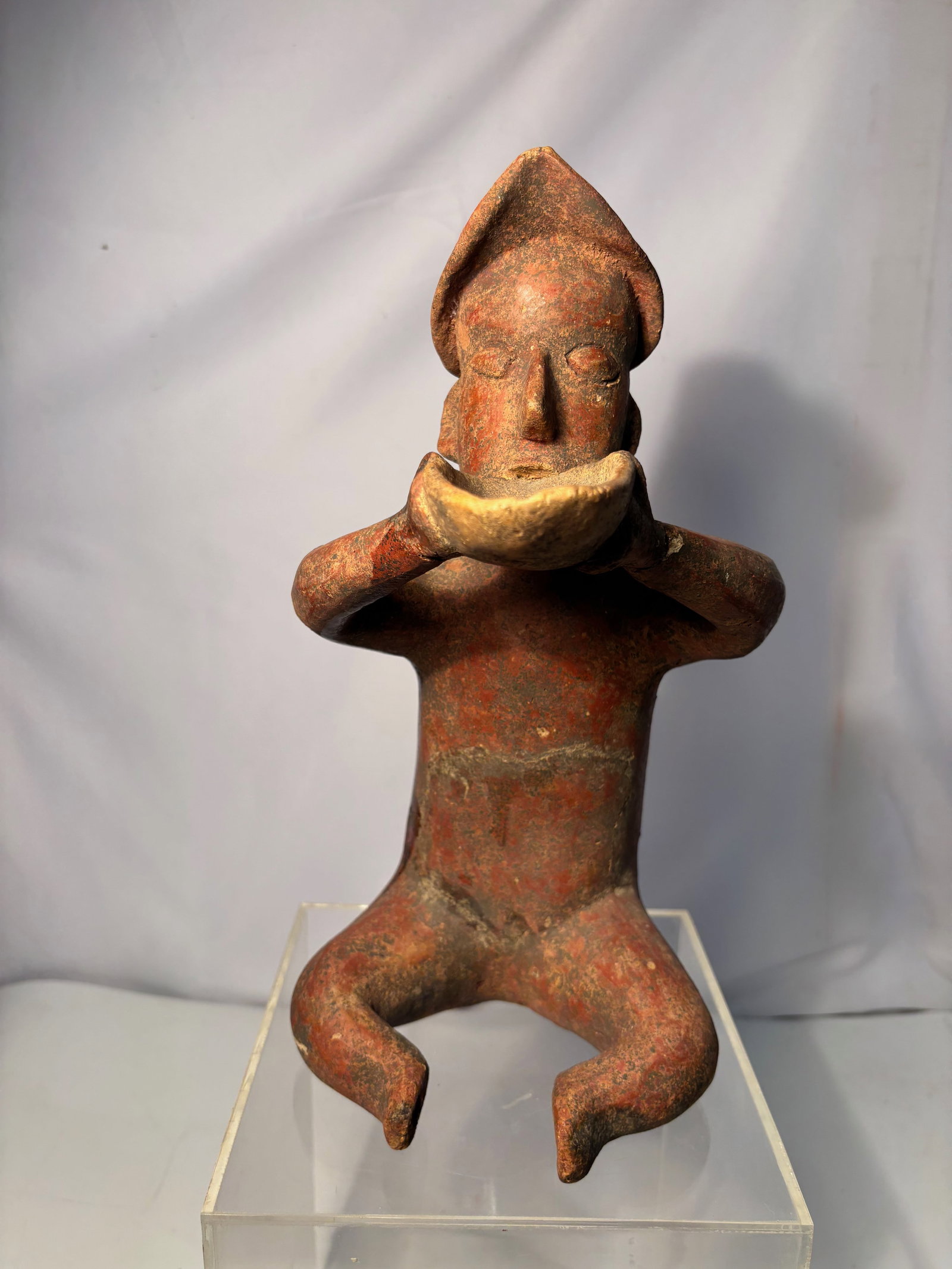 A Pre-Columbian Style Terracotta Seated Figure: Terracotta seated figure rendered in a Pre-Columbian style, depicting a stylized male form with bent legs, outstretched arms, and a pointed headdress. The figure features a red-brown surface with area