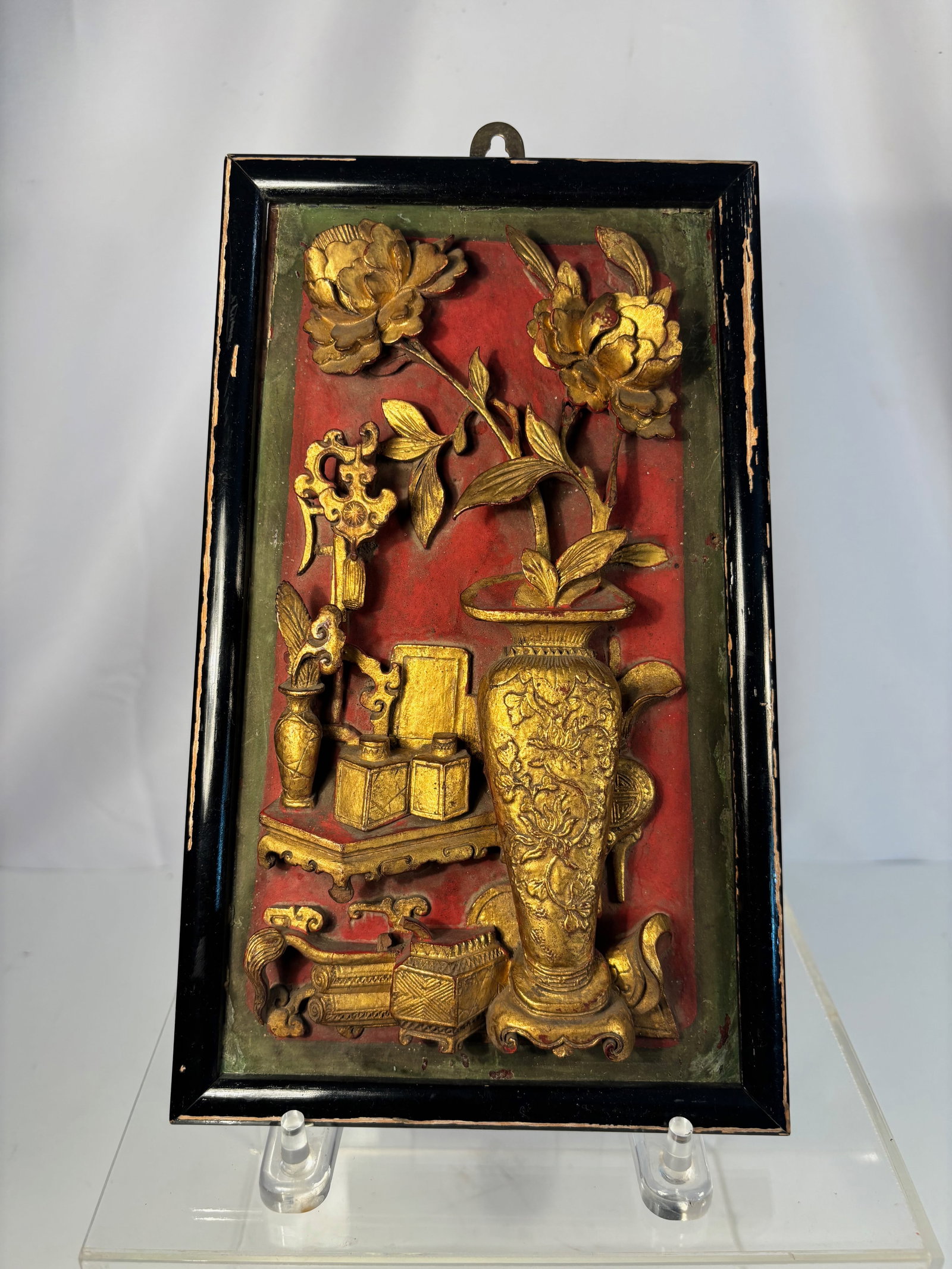 A Chinese Giltwood Carved Panel with Vases and Scholar’s Objects, Qing Dynasty: Qing dyansty(1644-1912); A finely carved and gilt wood panel (zhaoxiang ban) depicting an arrangement of vases with flowers, books, scrolls, and ritual objects, all set against a red lacquer ground an