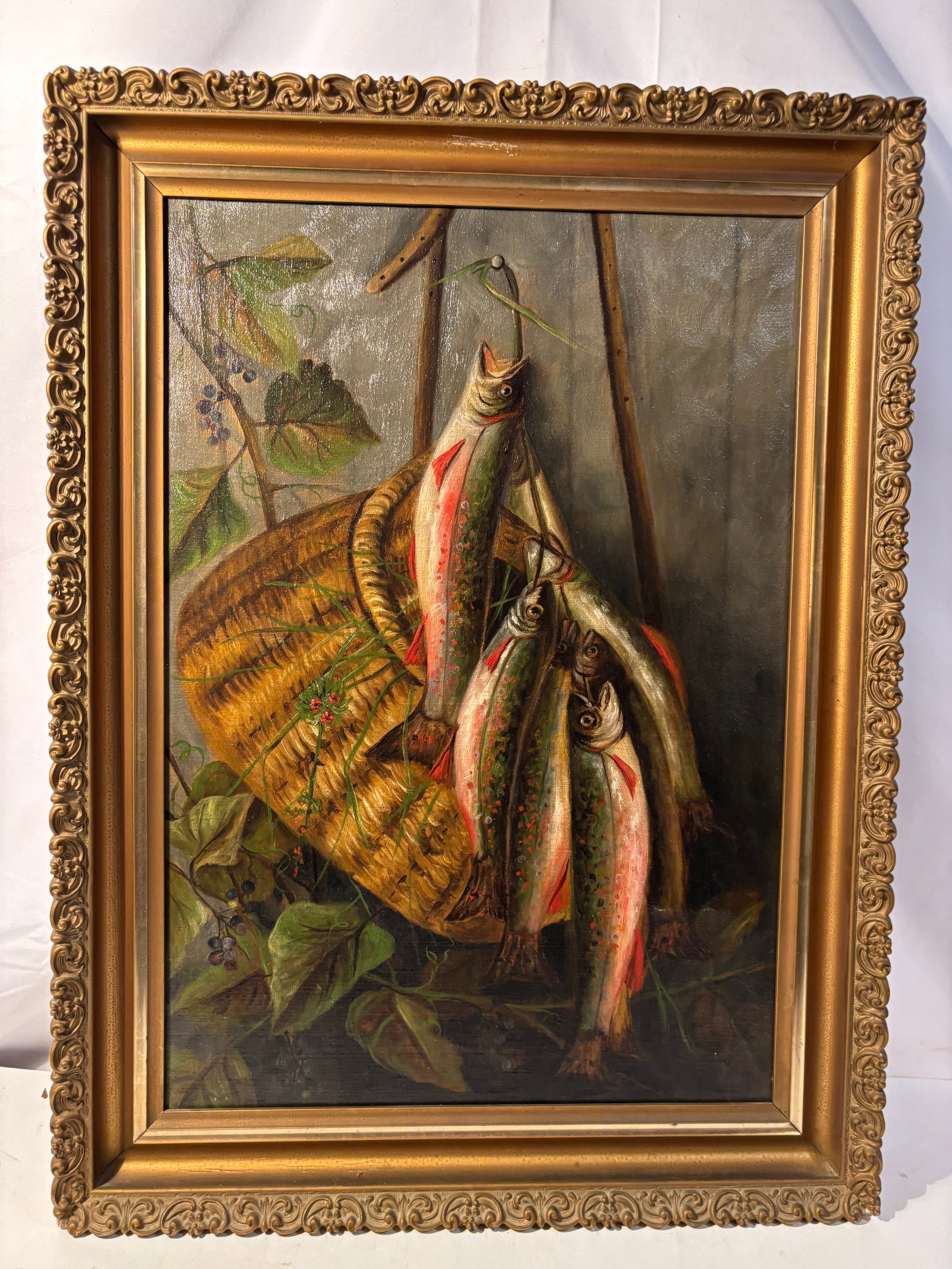 Signed Hanging Trout Still Life Oil Painting on Canvas, Framed 19th century: A signed oil painting on canvas depicting a rustic sporting still life with several freshly caught trout suspended from a fishing line above a woven creel basket, surrounded by leafy woodland foliage.