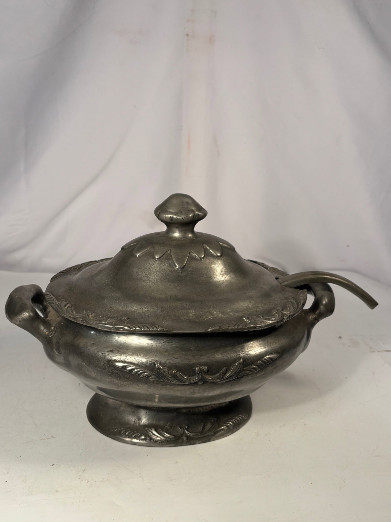 Vintage Pewter Covered Soup Tureen with Ladle: A decorative pewter soup tureen featuring an oval body, twin handles, raised foot, foliate relief decoration, and a domed lid topped with a fruit-form finial. Accompanied by a matching curved serving 