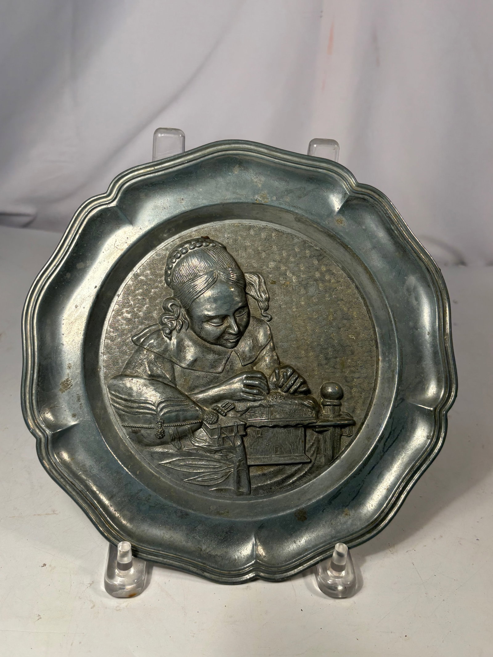Vintage German Zinn Pewter Lacemaker Wall Plate: A vintage German Zinn pewter wall plate featuring a raised relief scene of a young woman sewing or making lace at a table. Designed with a scalloped rim and fitted with a hanging tab. The reverse bear