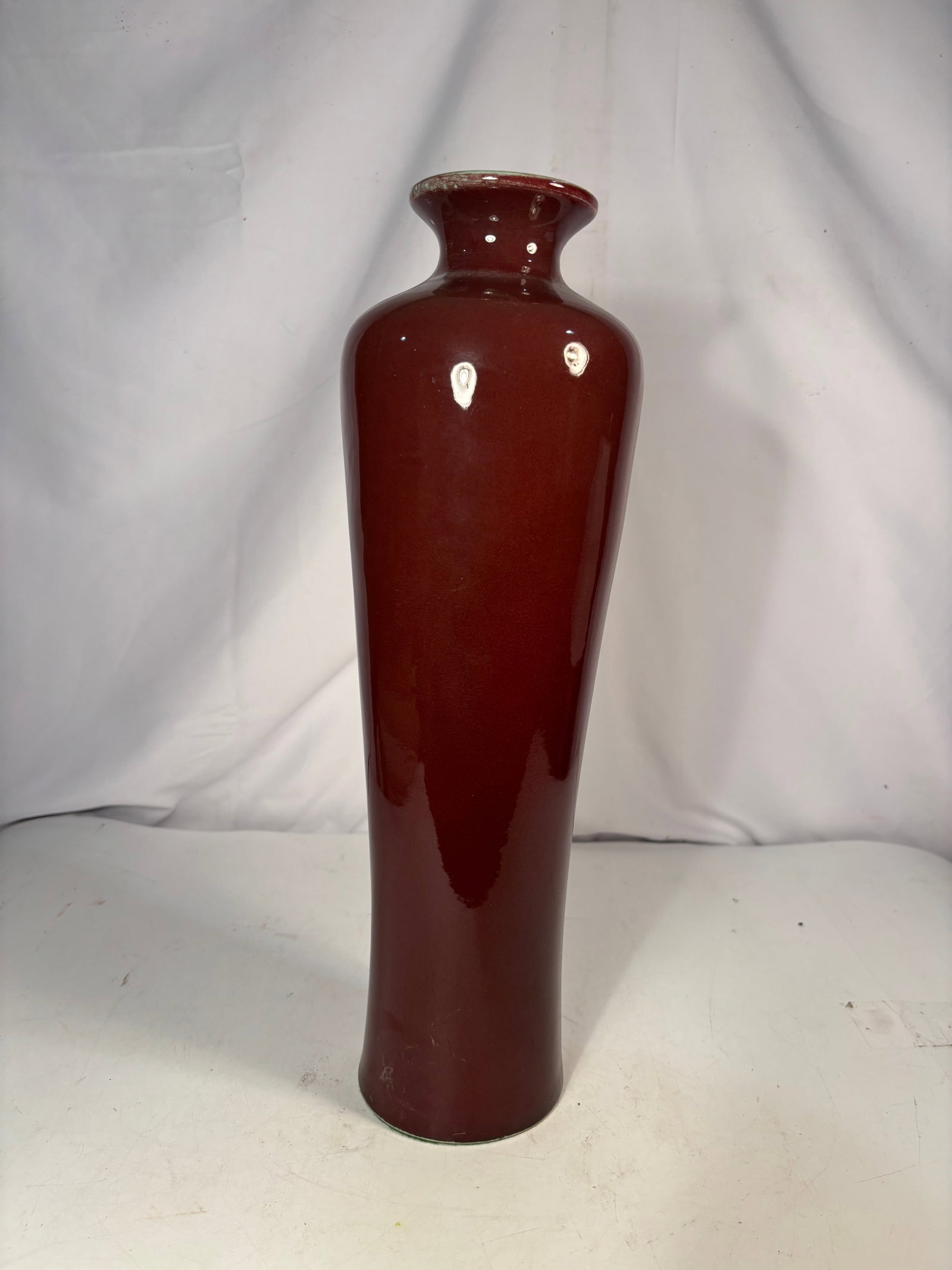 Chinese Oxblood-Glazed Ceramic Vase: A single Chinese ceramic vase with a tall tapered body, rounded shoulders, short neck, and flared rim, covered in a deep oxblood-red glaze. The underside is covered with green felt and bears a Chinese