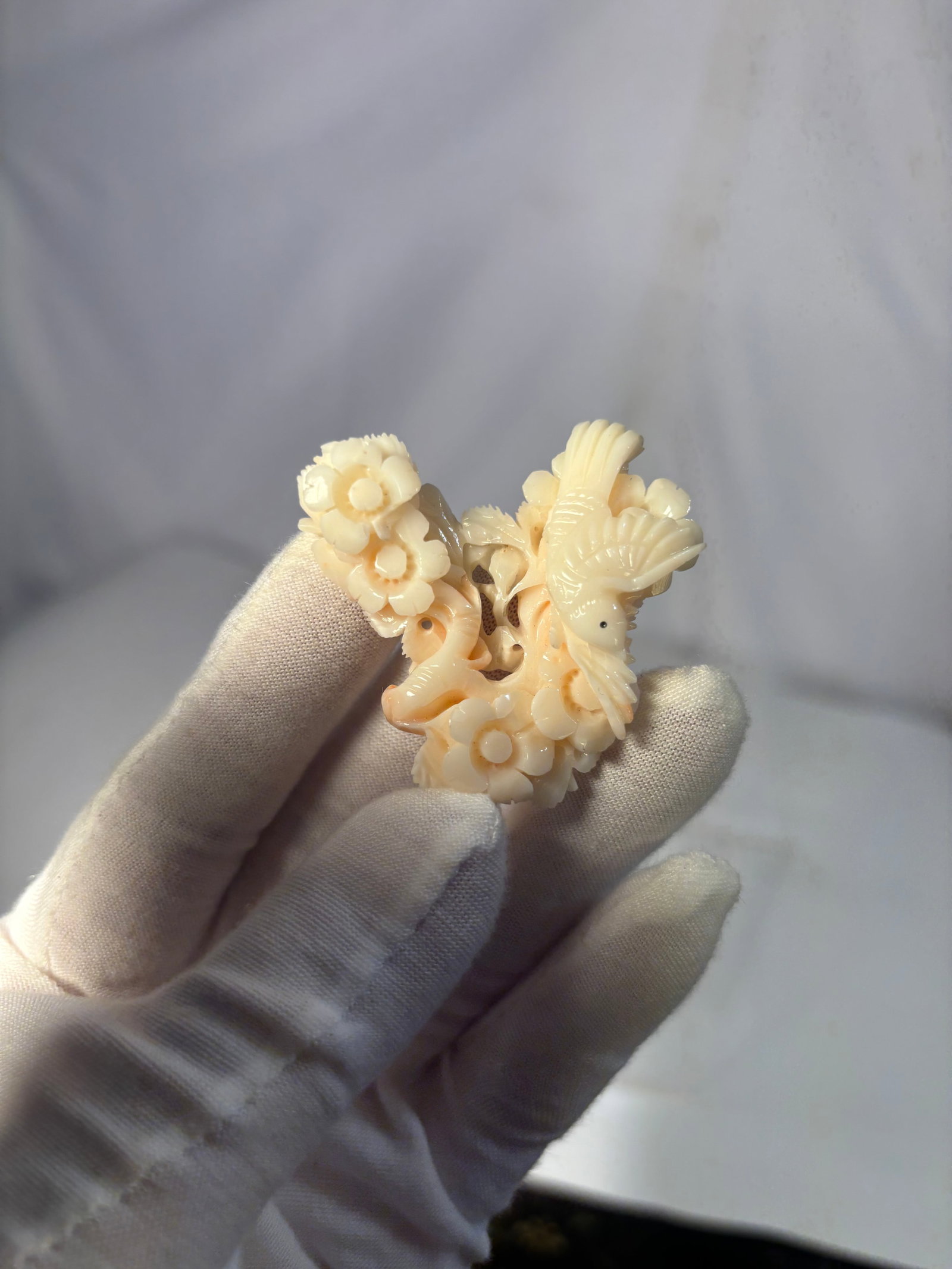 A Carved Pink Coral Floral and Bird Pendant: A finely carved pendant or small ornament in a pink coral featuring a delicate openwork design of birds among blossoming flowers. The carving displays a soft palette of pale pink, cream, and warm ivor