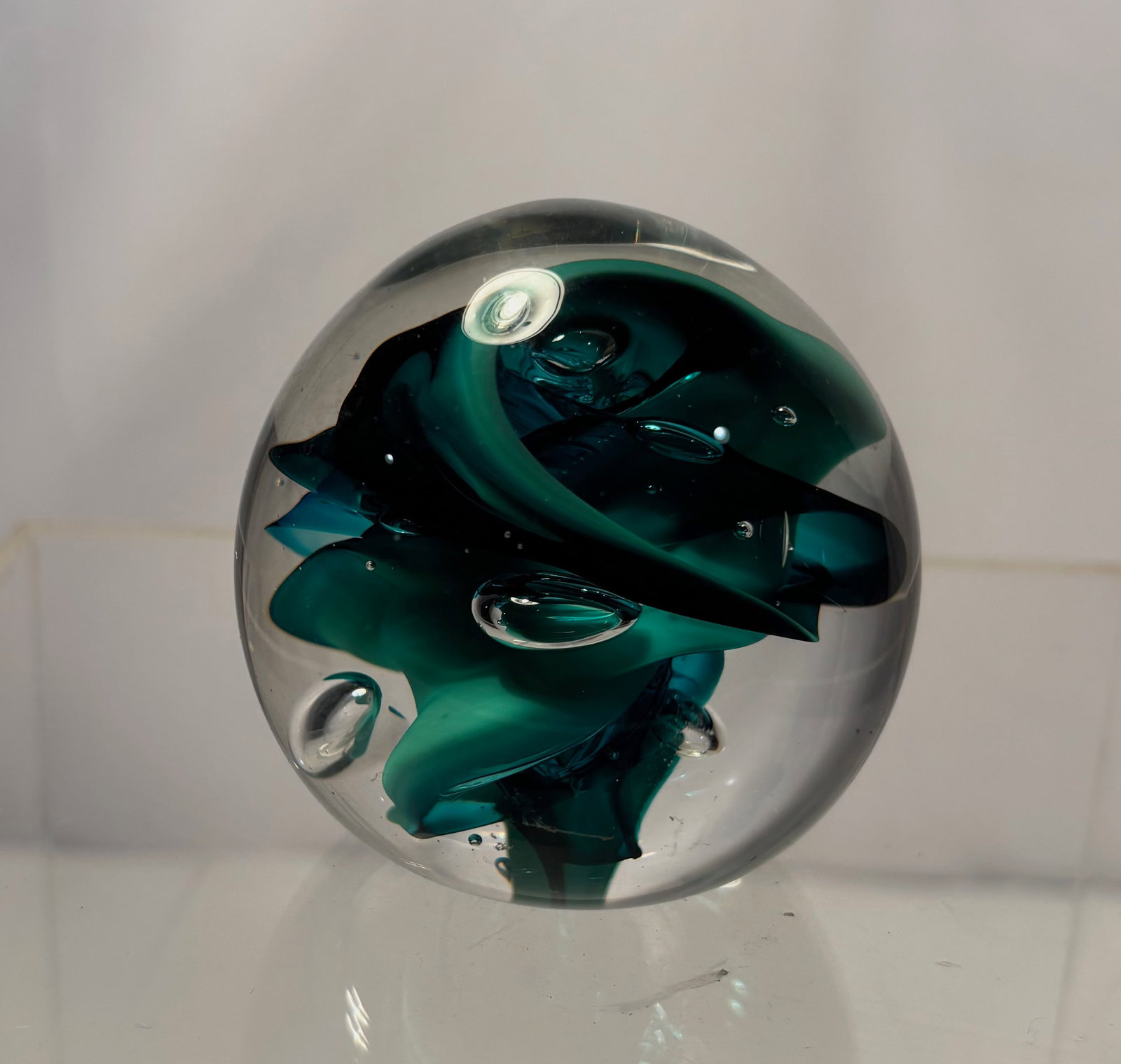 A Signed Studio Art Glass Teal & Green Vortex Paperweight: A studio art glass paperweight featuring a dynamic internal teal and green vortex form encased in clear glass. The abstract ribbon-like inclusions create strong depth and movement, enhanced by control