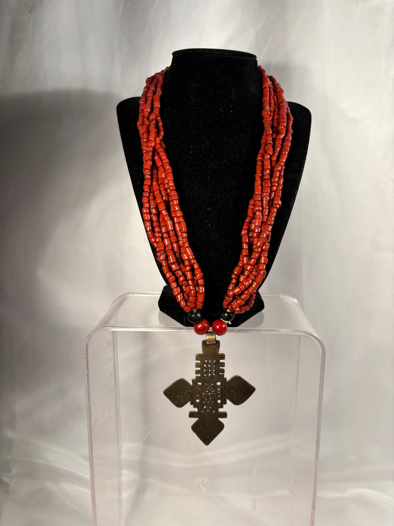 Vintage Ethiopian Multi-Strand Coral Necklace with Coptic Cross Pendant: A vintage Ethiopian necklace composed of multiple strands of small, irregular red coral barrel beads. The center is accented by four larger round glass beads—two red and two black—leading to a pie